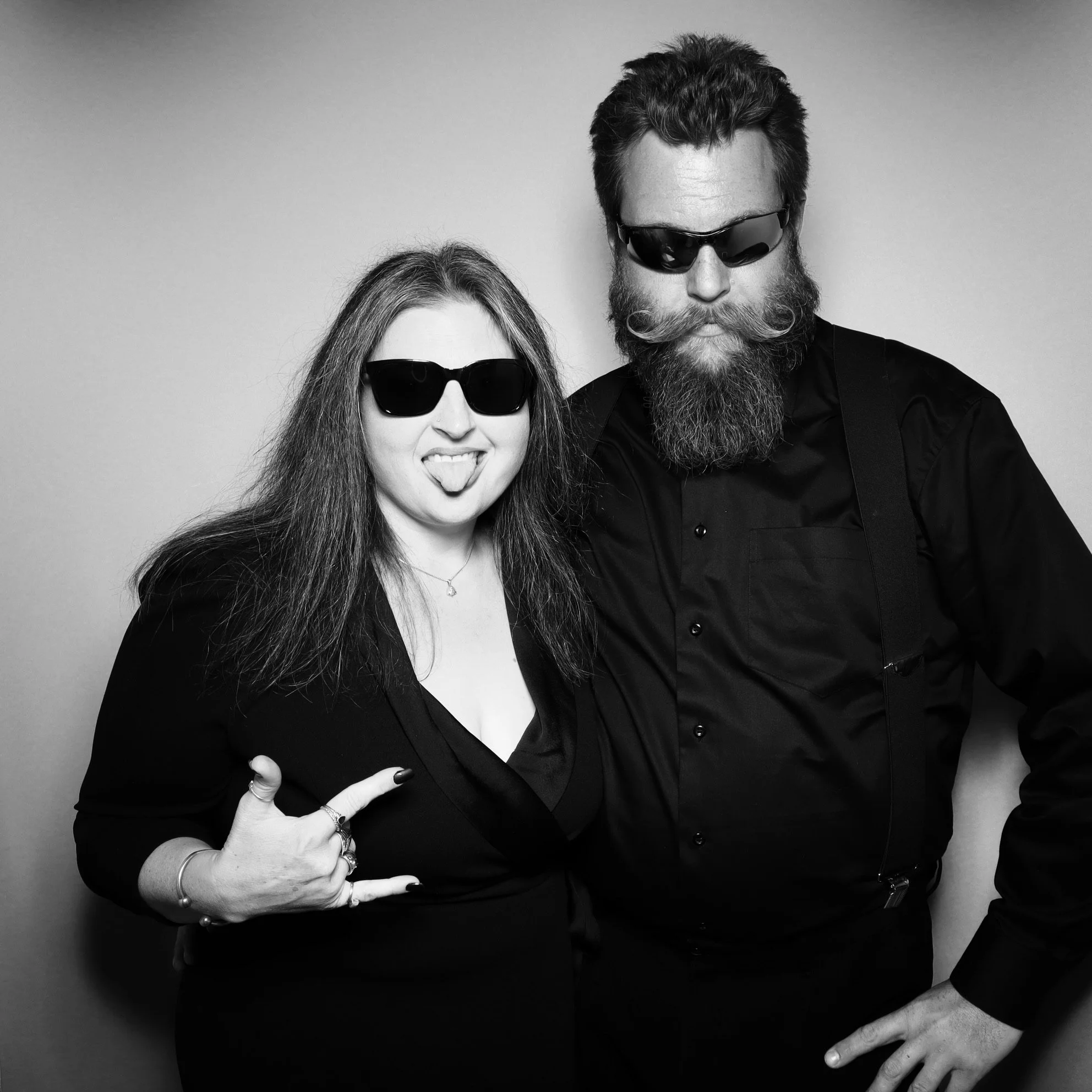Black and white photo of a woman and man wearing sunglasses, making rock and roll hand signs, with the woman sticking out her tongue and the man with a beard and mustache.