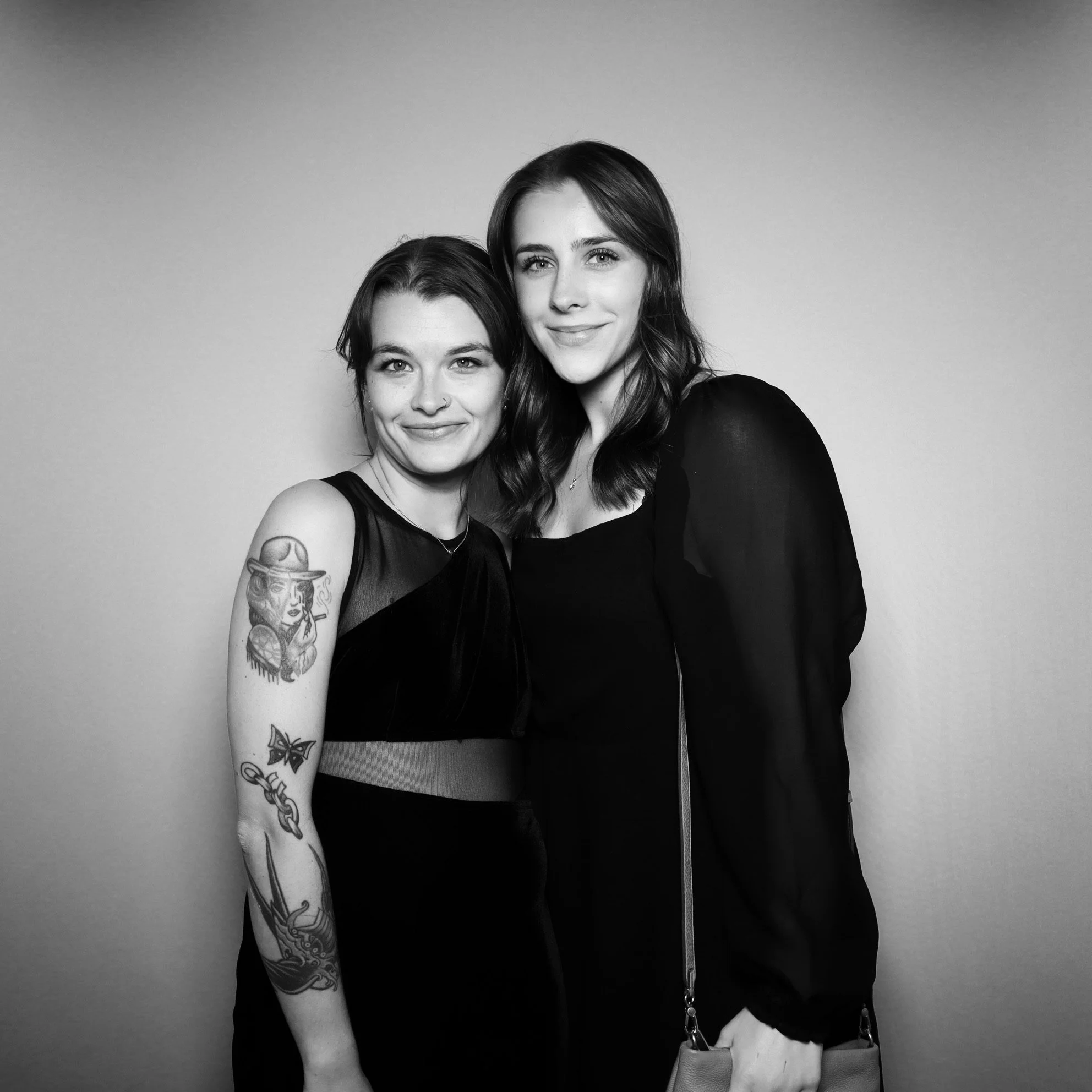 Two women in black dresses standing close together, smiling, against a plain background.