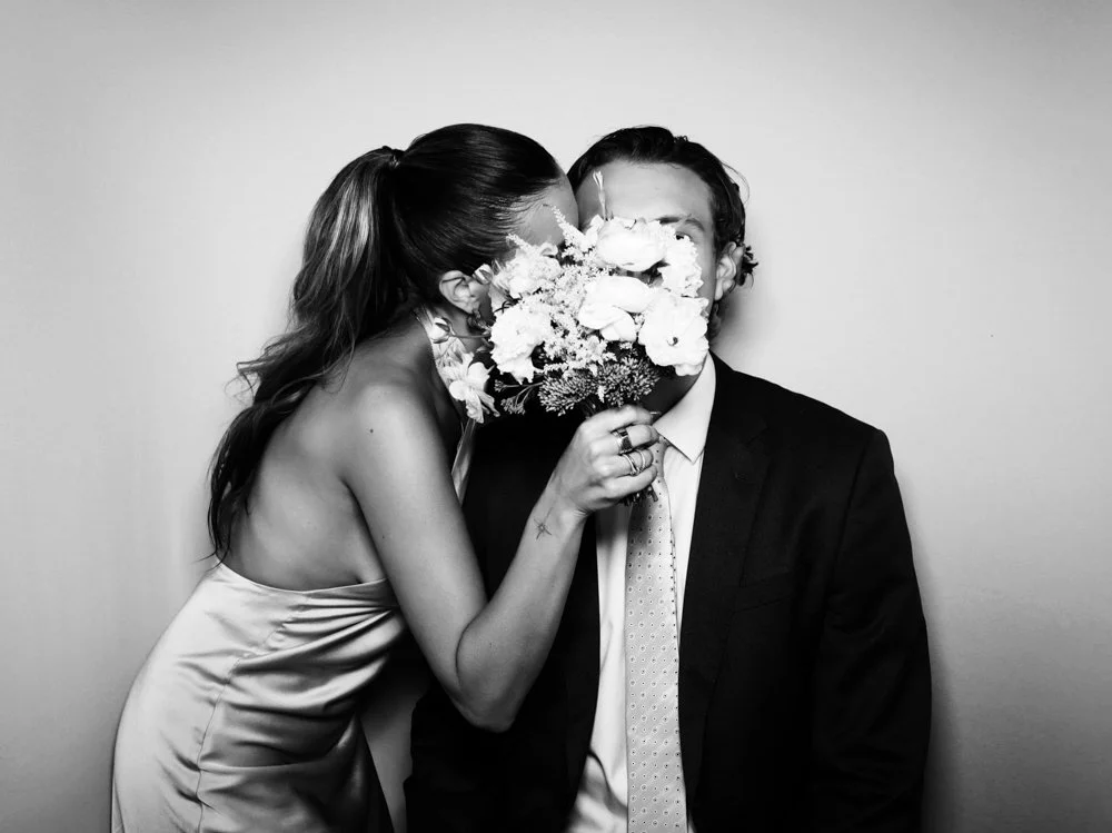 A woman in a satin dress leans in to kiss a man in a suit on the cheek while holding a bouquet of flowers in front of his face.