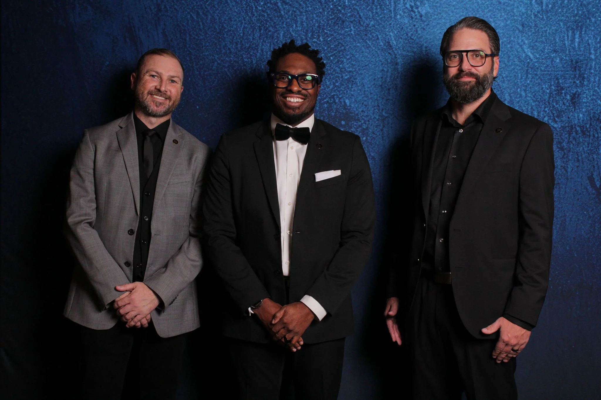 Three men in formal suits standing against a dark blue textured background.