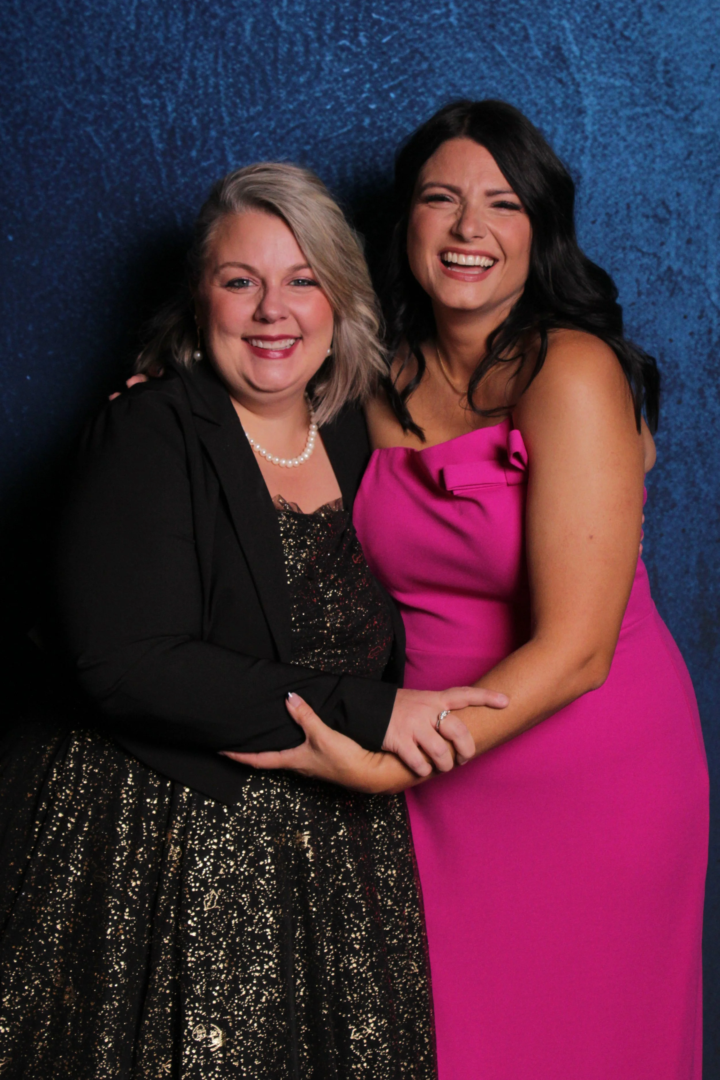 Two women smiling and embracing against a blue background, one wearing a black dress with sparkles and pearl necklace, the other in a pink strapless dress.