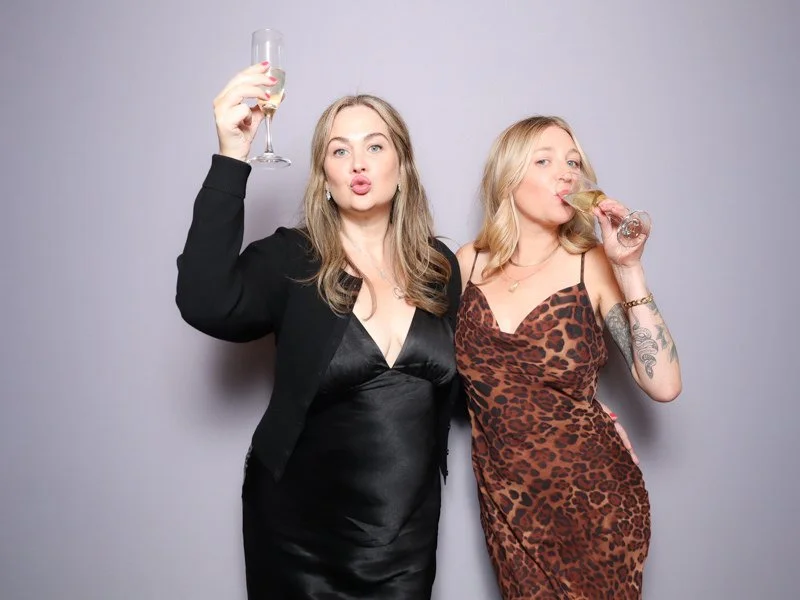 Two women celebrating with champagne and wine glasses, one in a black dress, the other in a leopard print dress, against a plain gray background.