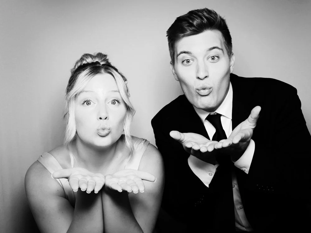A woman and man in formal attire blowing kisses towards the camera, both with playful expressions in a black-and-white photo.
