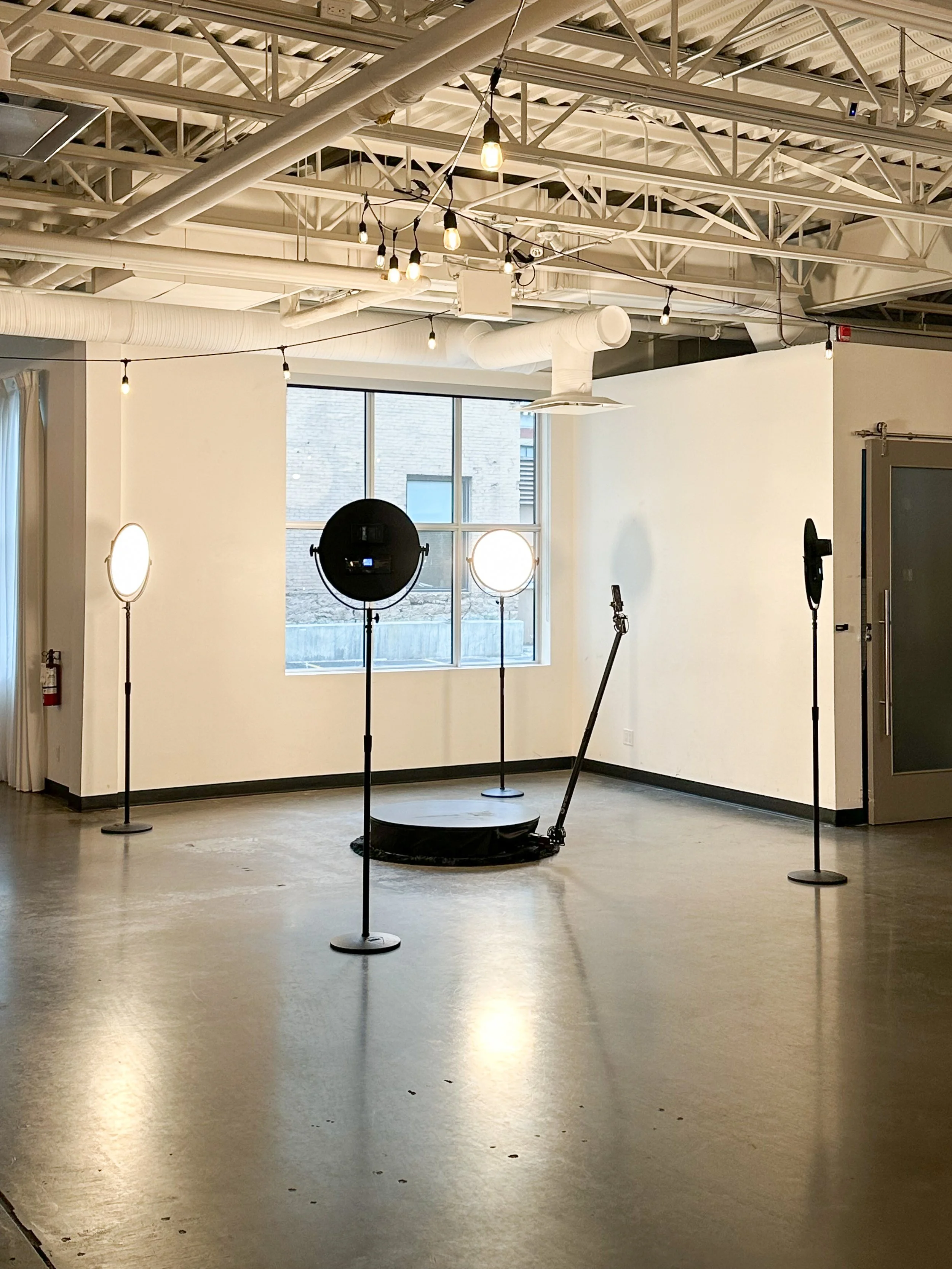 Empty photography or video studio with four standing light stands and large diffused lights, a black circular platform, and a brown roller door.