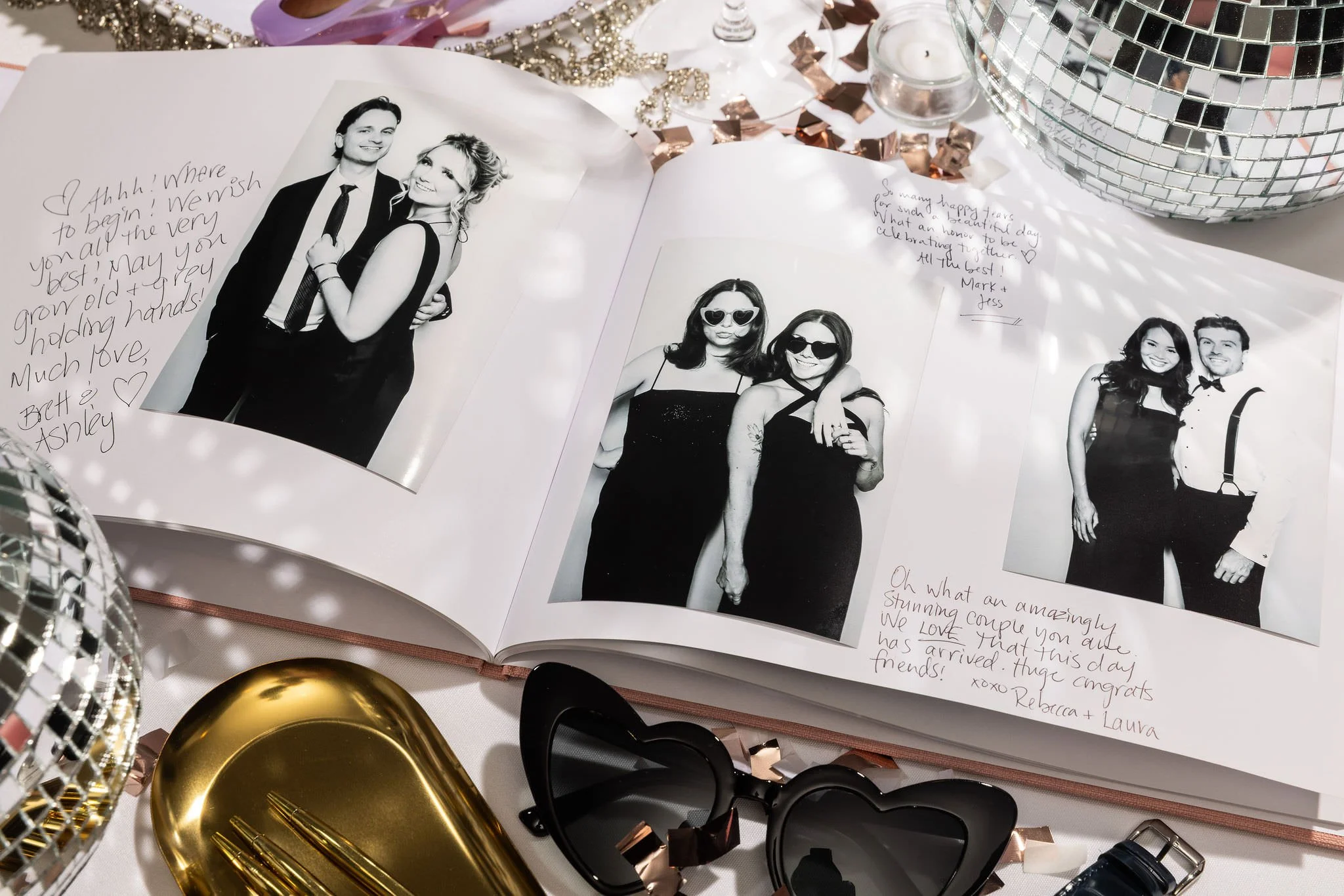 Open photo album with black-and-white wedding photos and handwritten notes, surrounded by party decorations including a disco ball, gold tray, heart-shaped sunglasses, and a tape measure.