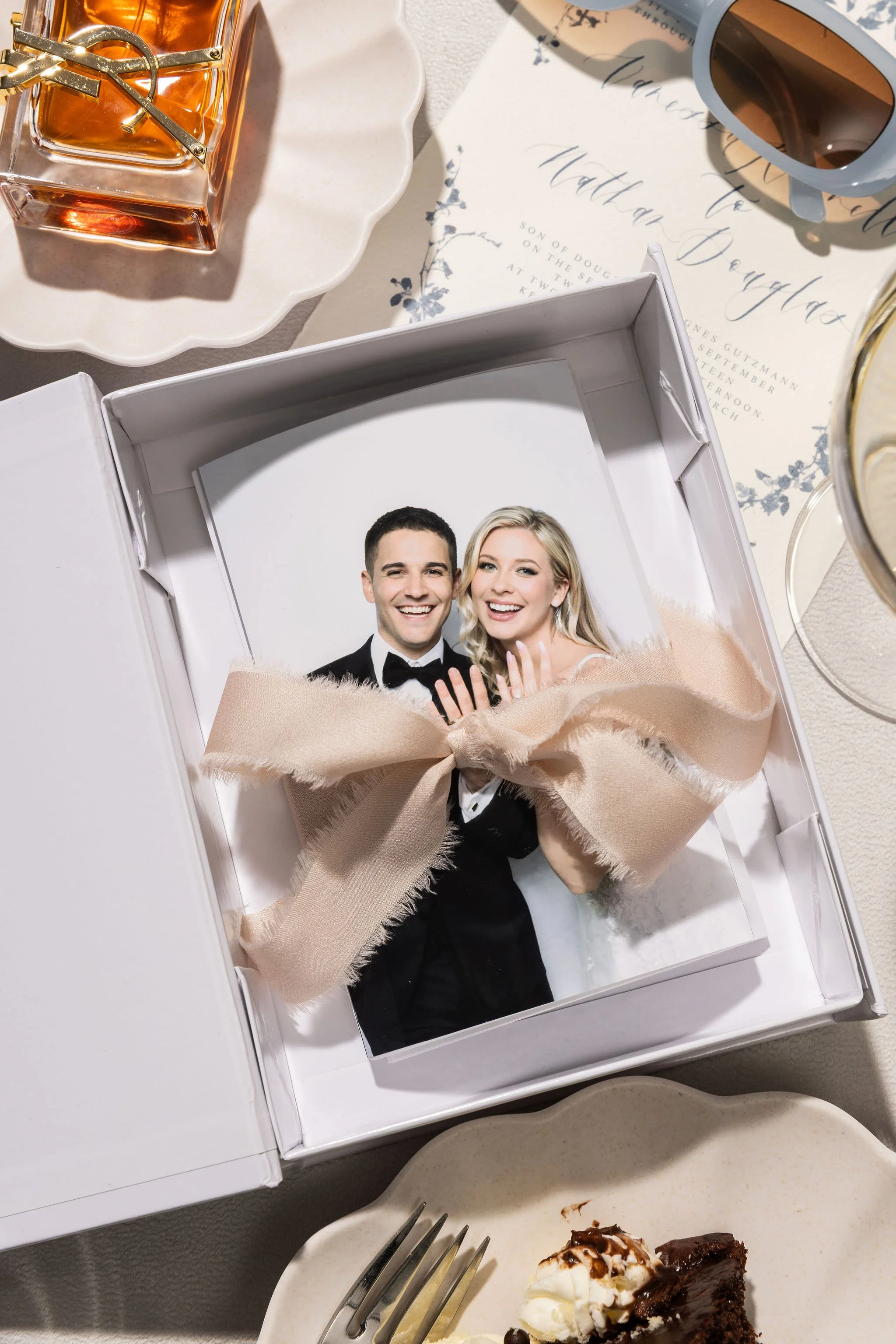 A wedding photo of a happy couple in a box on a table, surrounded by dessert plates, sunglasses, and a handwritten wedding invitation.