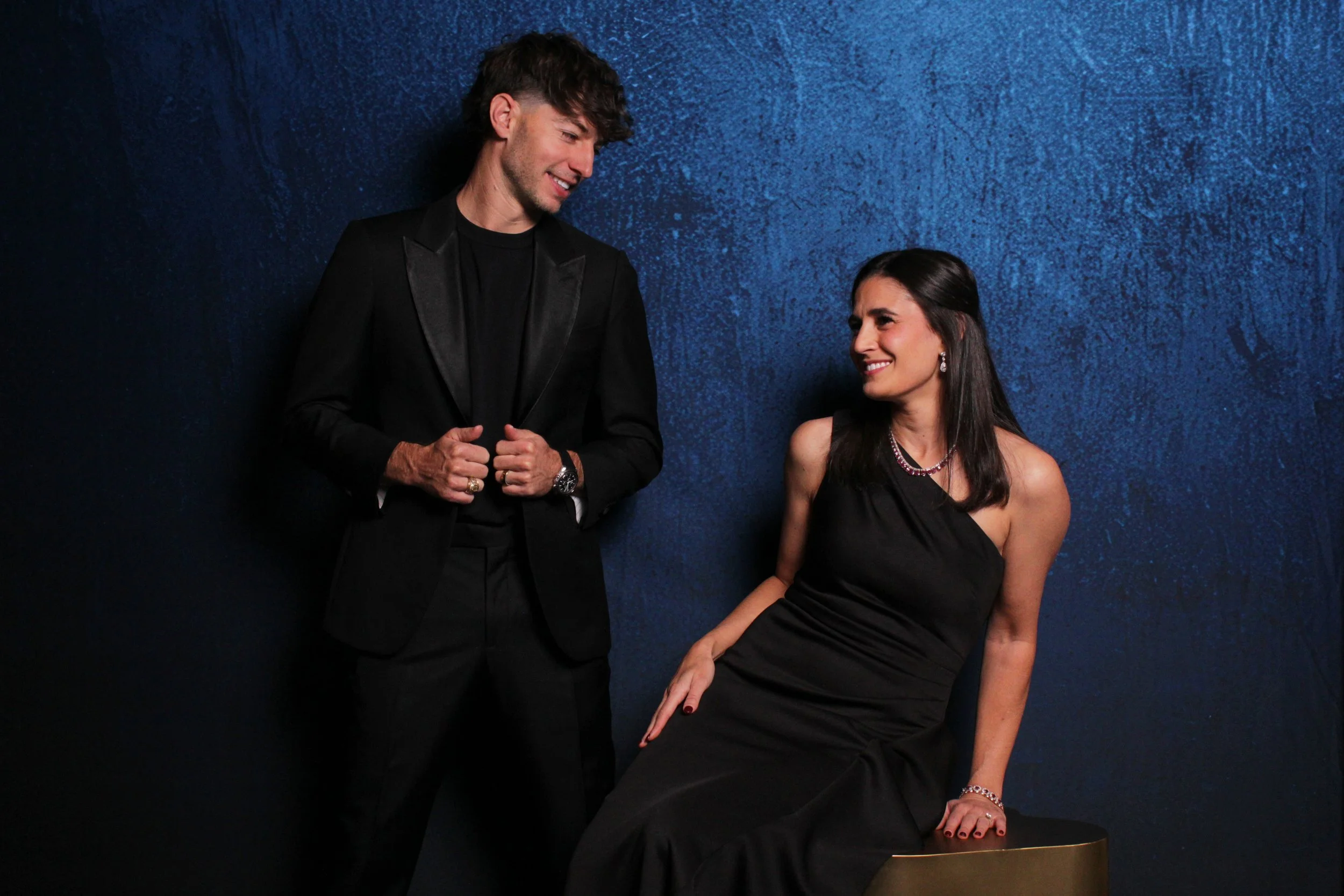 A man in a black tuxedo and a woman in a black evening gown smiling at each other against a dark blue textured wall.