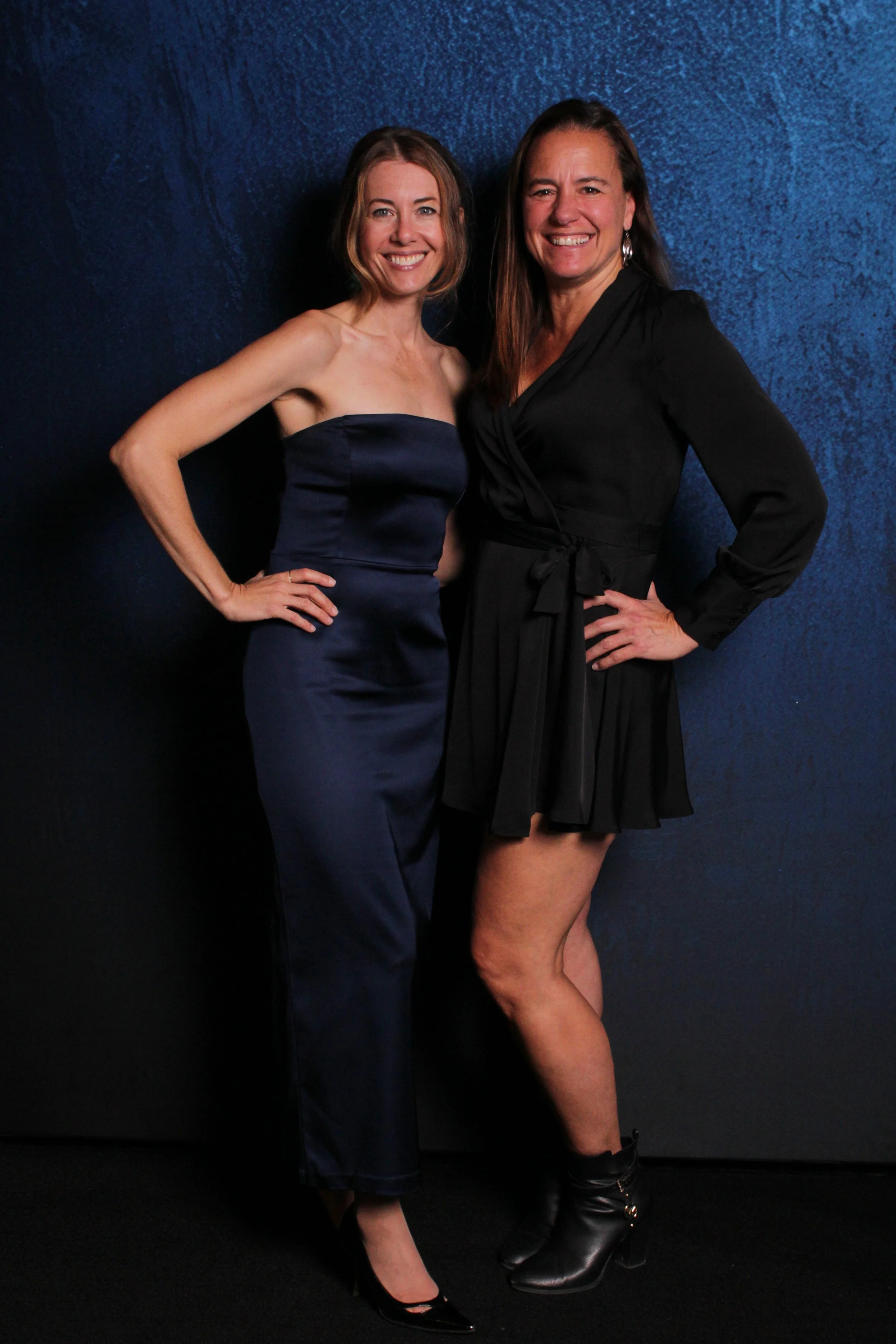 Two women smiling and posing together against a dark blue textured background, one wearing a strapless navy blue dress and the other in a black wrap dress with long sleeves and high heels.