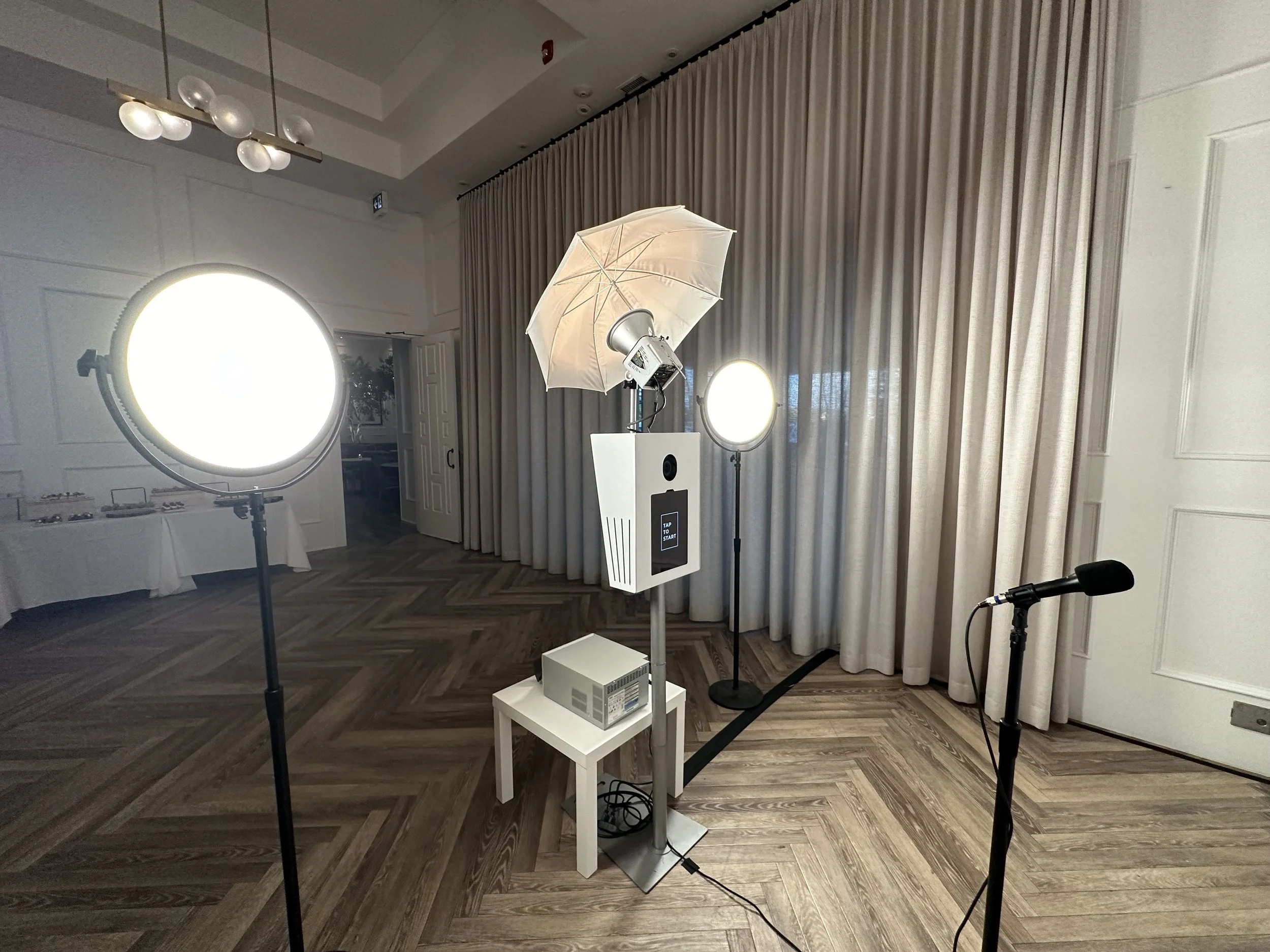 Photo of a professional photography or video setup with large studio lights, reflectors, a white umbrella, and a microphone in a room with wooden flooring and beige curtains.