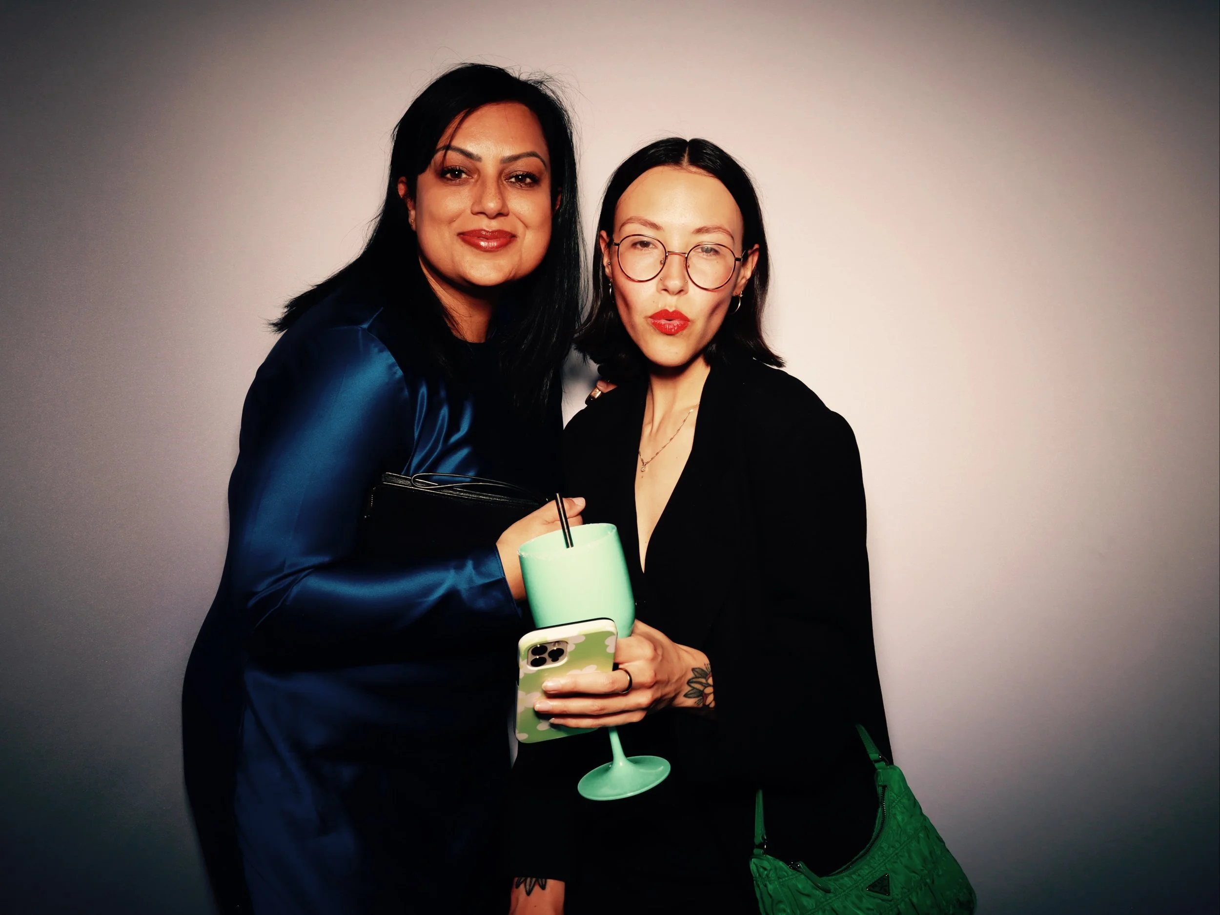 Two women standing close together, one with black hair wearing a blue satin jacket, the other with glasses and tattoos, holding a green cocktail and a phone, against a plain background.