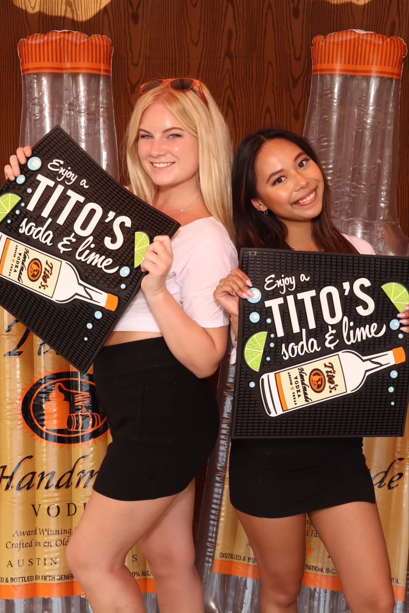 Two young women holding signs, smiling, in front of a backdrop with large inflatable Tito's vodka bottles. The signs read 'Enjoy a Tito's soda & lime.'