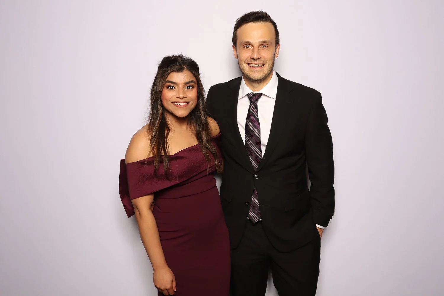A woman in a burgundy off-shoulder dress and a man in a black suit with a striped tie, standing together against a plain light background, smiling for the camera.