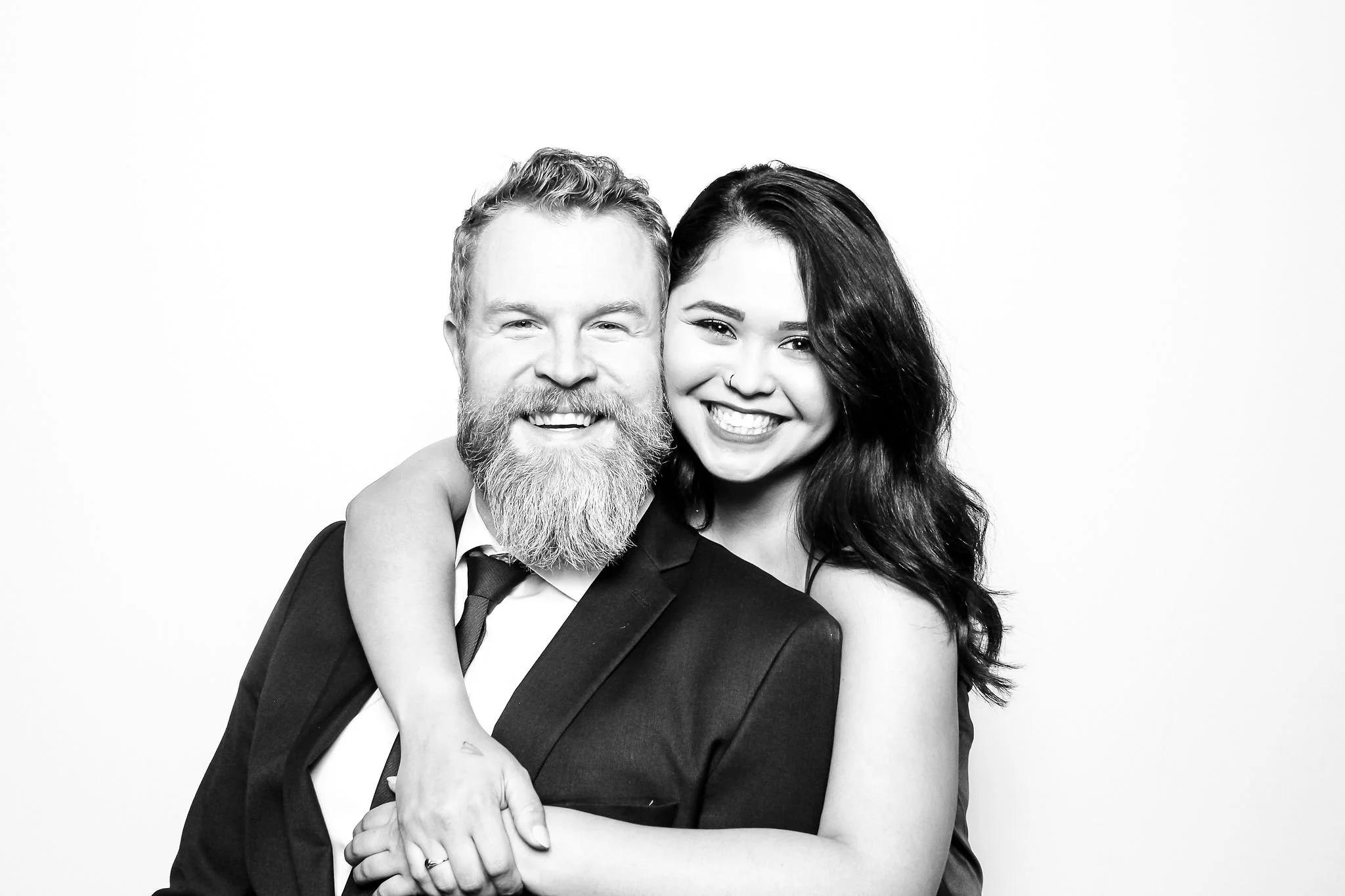 Black and white photo of a smiling man with a beard and mustache dressed in a suit, being embraced by a young woman with long dark hair, smiling, with her arm around his shoulders, against a plain white background.