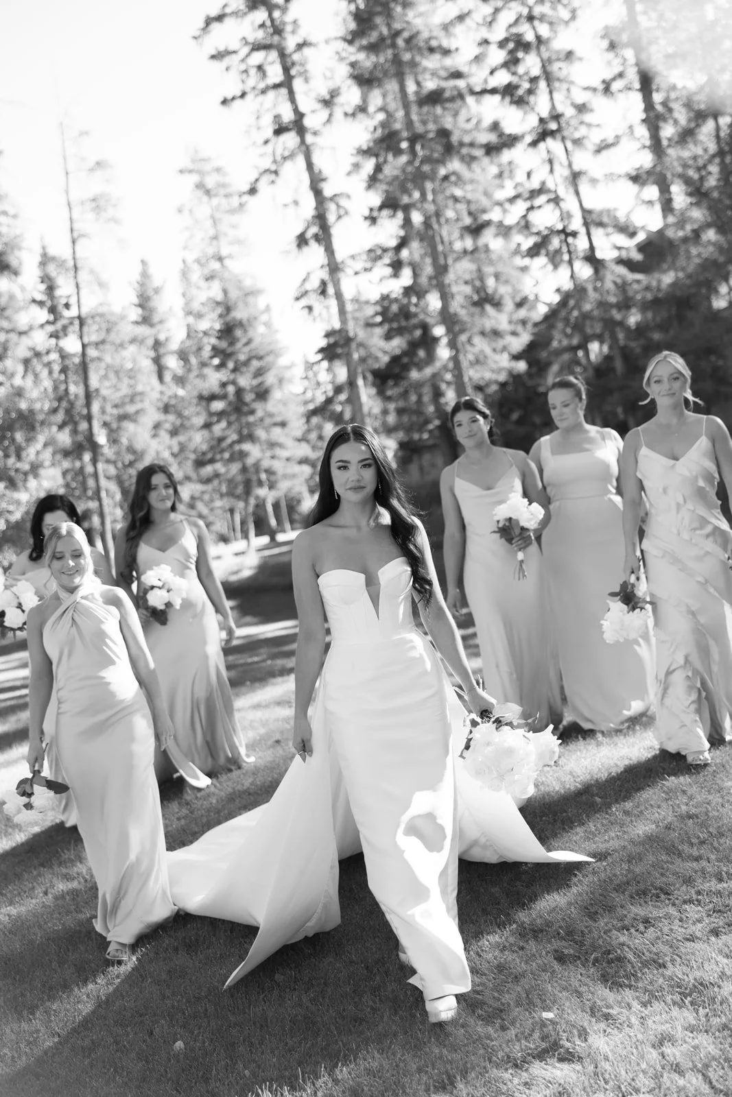 Black and white photo of a bridal procession outdoors, with women in elegant dresses walking on a grassy area among tall trees, led by a bride in a strapless wedding gown holding a bouquet.
