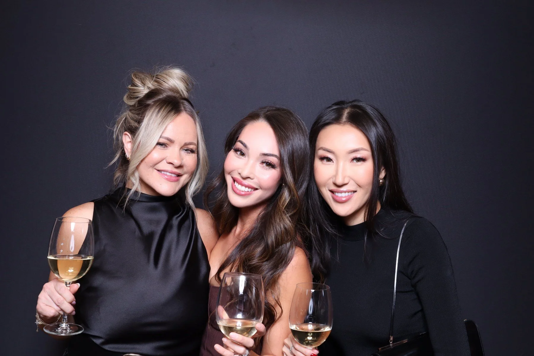 Three women smiling and holding glasses of white wine, posing against a dark background.
