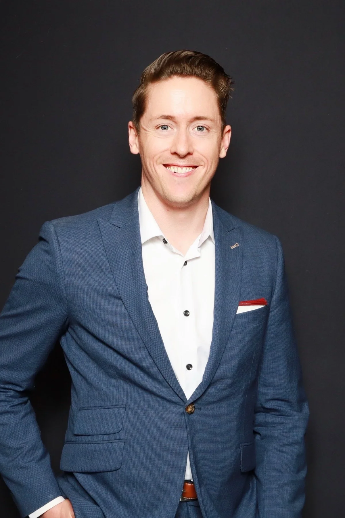 Portrait of a young man with light skin, brown hair, and blue eyes, dressed in a blue suit jacket and white shirt, smiling against a black background.