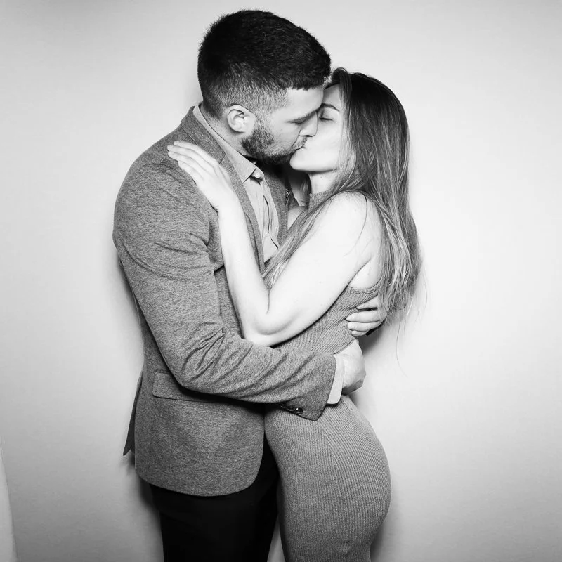 Black and white photo of a couple kissing, with the man in a blazer and the woman in a sleeveless dress, hugging each other against a plain background.