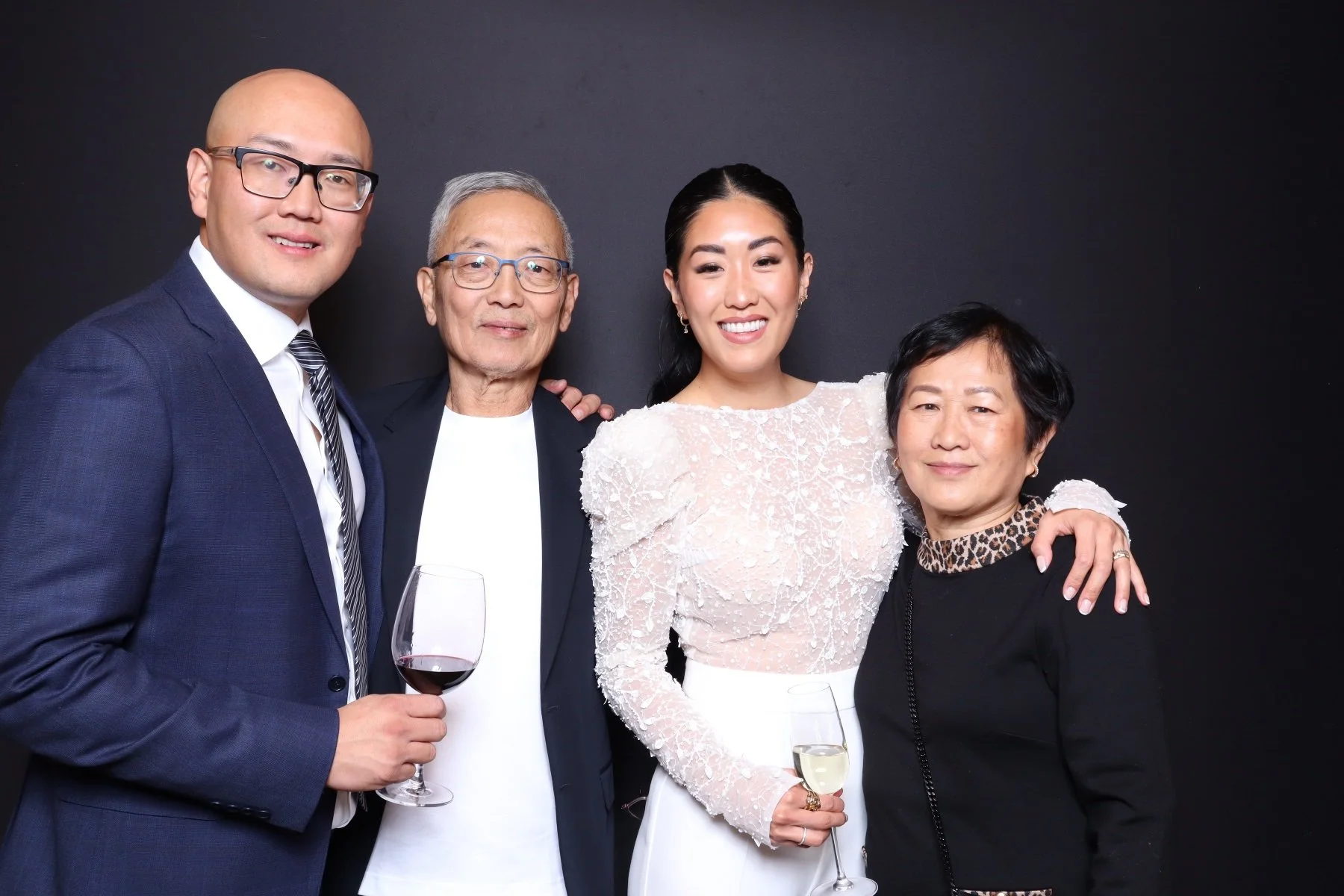 Four people at a celebration, holding glasses of wine, smiling against a black background.