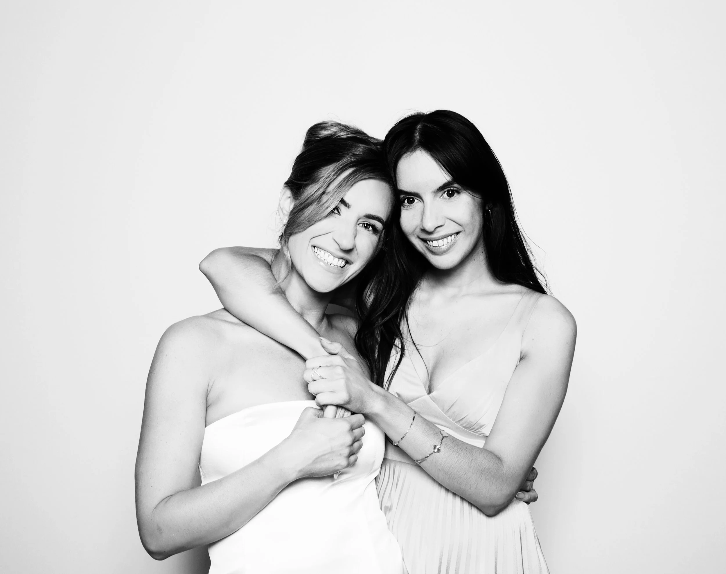 Two women standing close together, one winking and smiling, the other smiling, with the woman on the left wearing a strapless top and the woman on the right wearing a sleeveless dress.