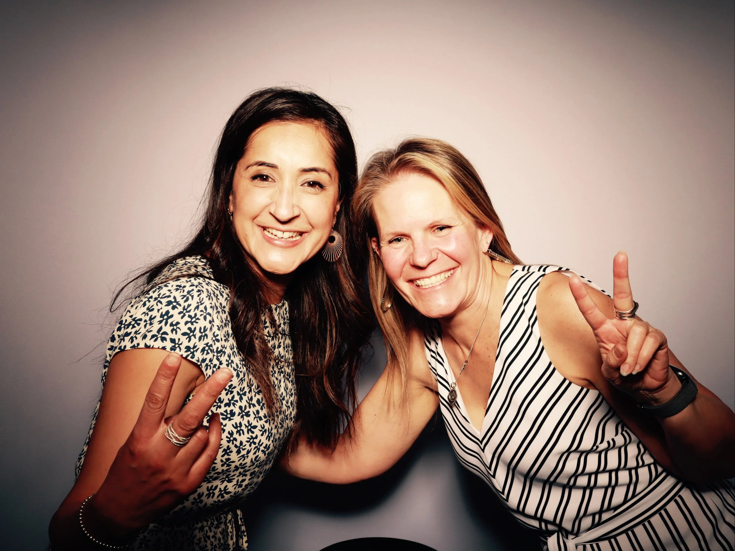 Two women smiling and making peace signs at the camera, wearing striped and patterned tops against a plain background.