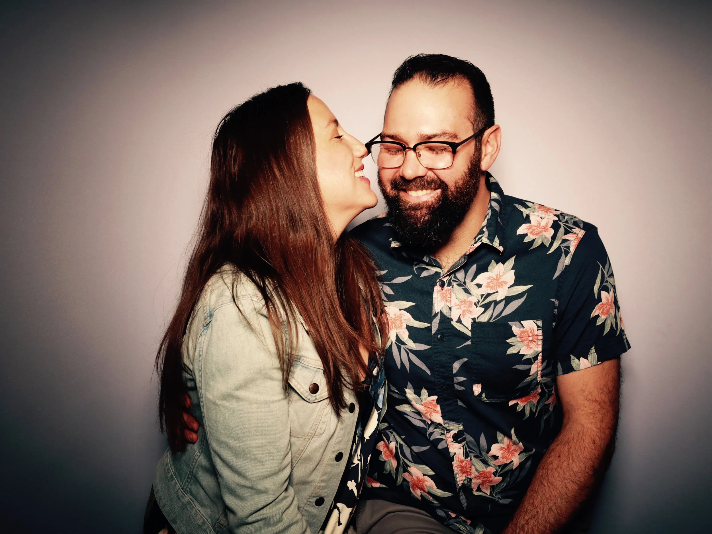 A couple sharing a joyful moment, with the woman leaning in to whisper or kiss the man, who is smiling and wearing glasses and a floral shirt, against a plain background.