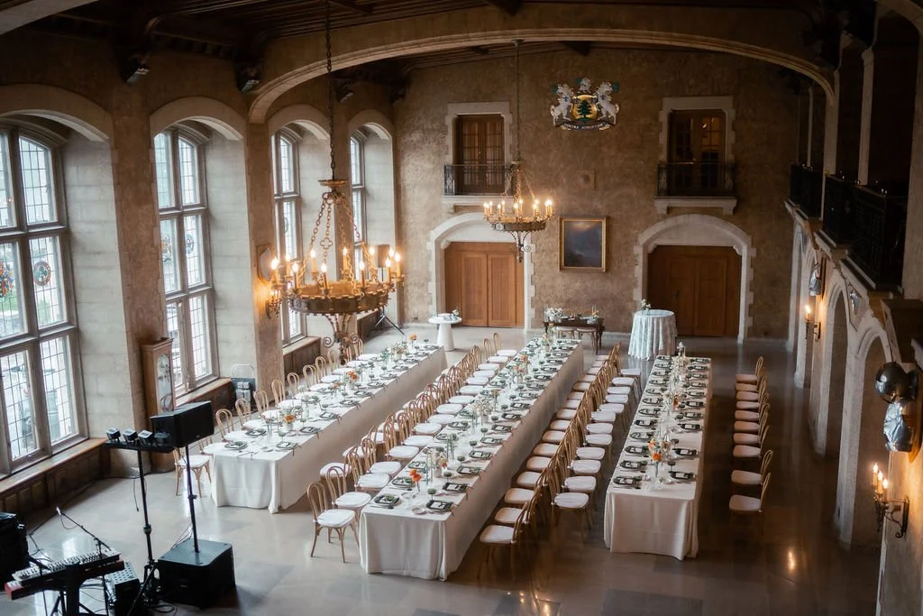 Elegant banquet hall with long dining tables covered in white tablecloths, set with plates, glasses, and cutlery, surrounded by wooden chairs with white cushions, large arched windows, and ornate chandeliers hanging from a high ceiling, decorated wit