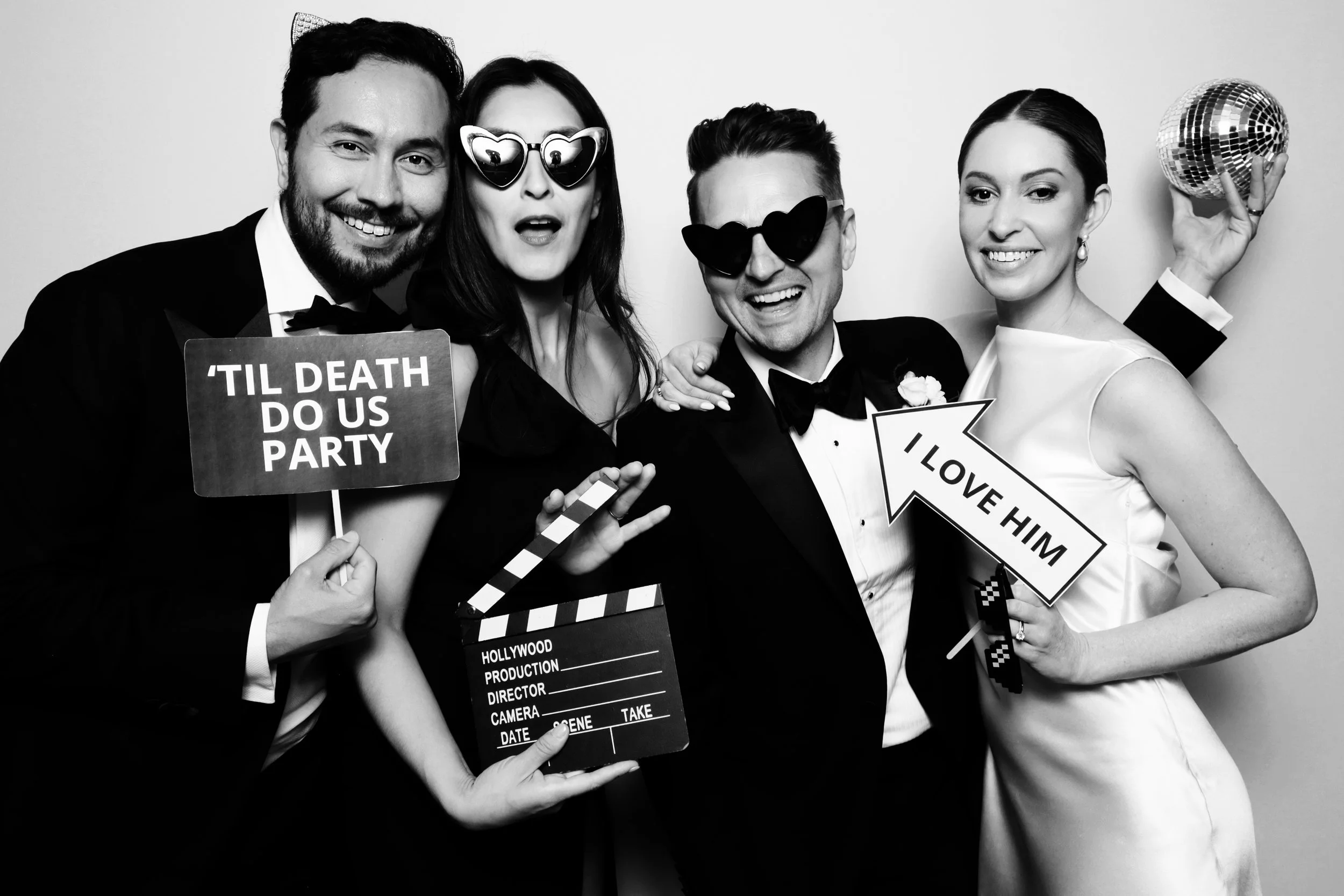 Group of five people celebrating at a party, dressed in formal attire, wearing sunglasses, holding signs that say 'Til death do us party', 'I love him', and a film clapboard, with one person holding a disco ball.