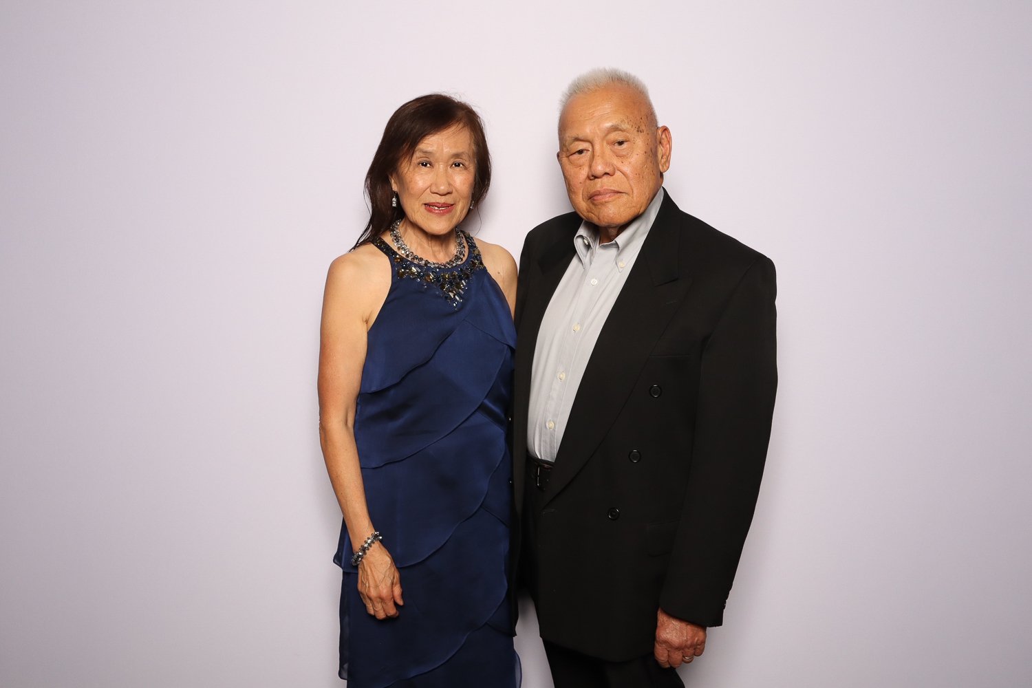 An elderly Asian couple dressed in formal attire, standing close together against a plain white background. The woman is wearing a navy blue evening gown with jewelry, and the man is in a black suit with a white shirt.