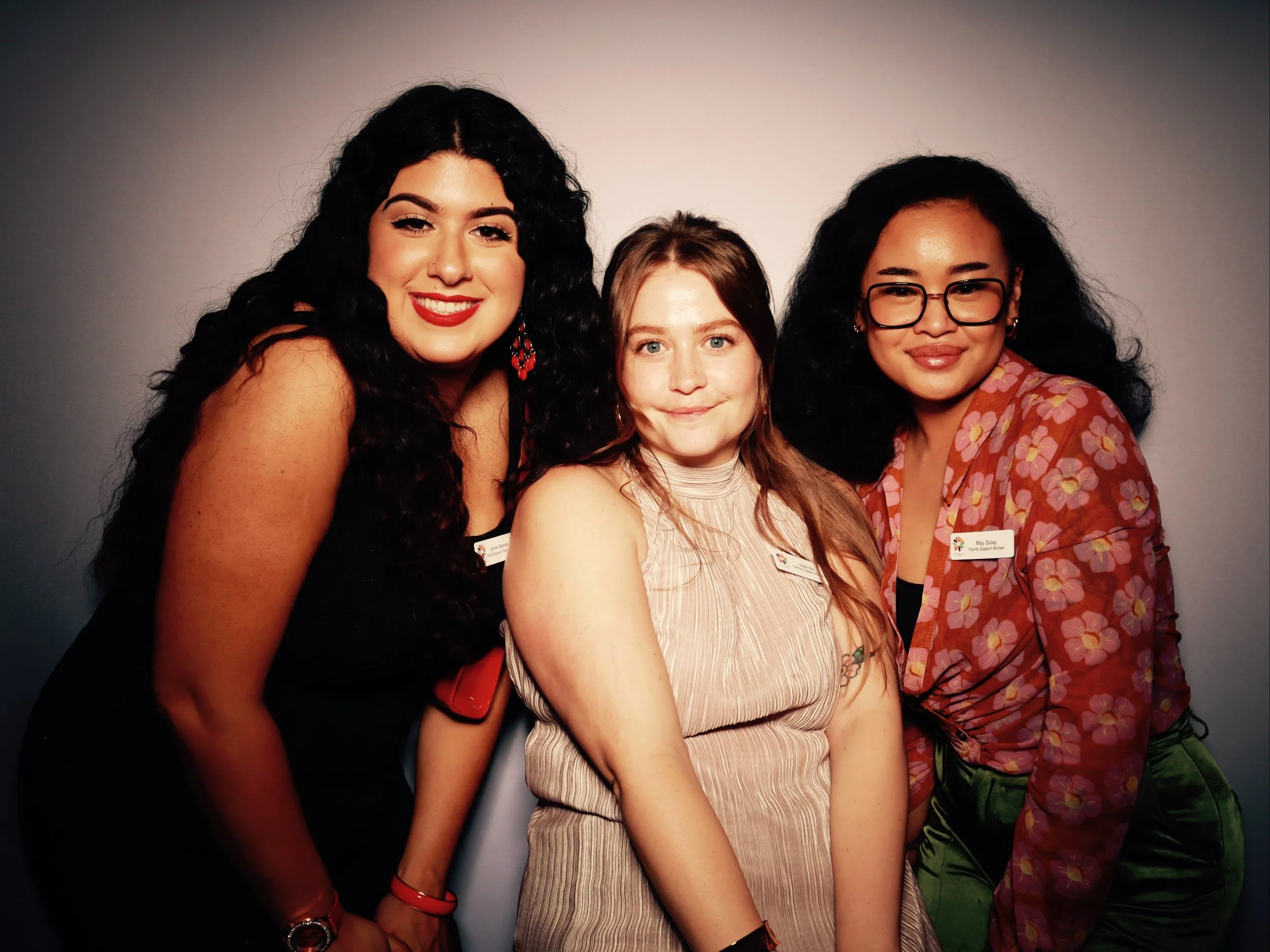 Three women standing together and smiling at the camera, wearing name tags, against a plain dark background.