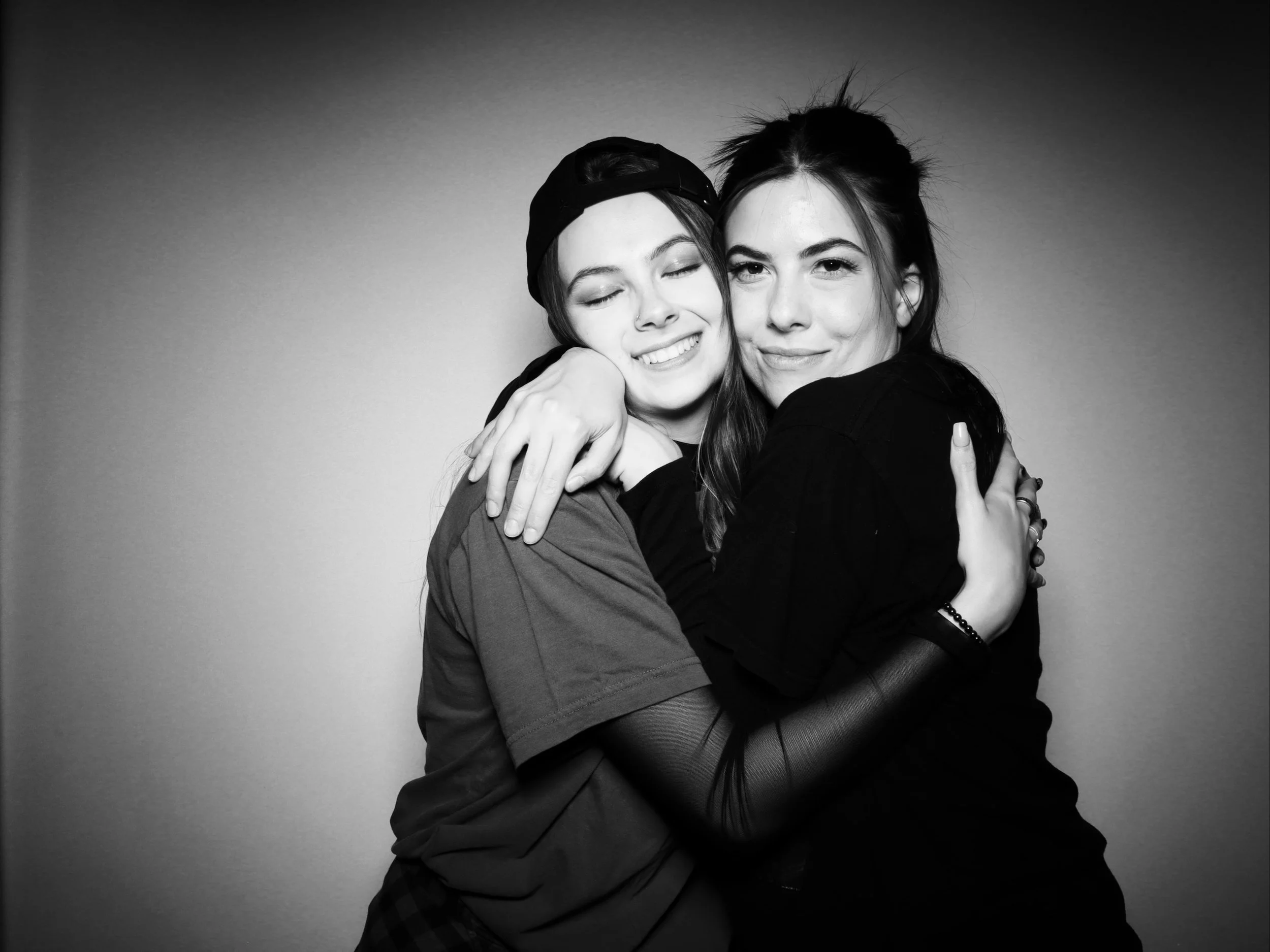 Two young women hugging each other in black and white. One has her eyes closed and is smiling, the other looks directly at the camera and is smiling slightly.