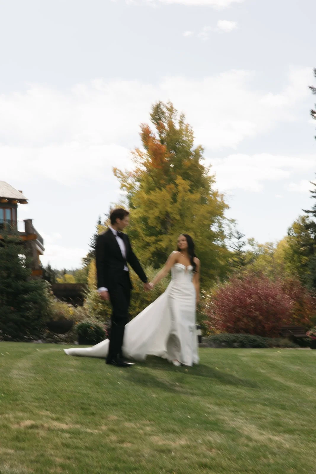 A bride and groom holding hands in a garden with autumn foliage, the bride in a white wedding gown and the groom in a black tuxedo.