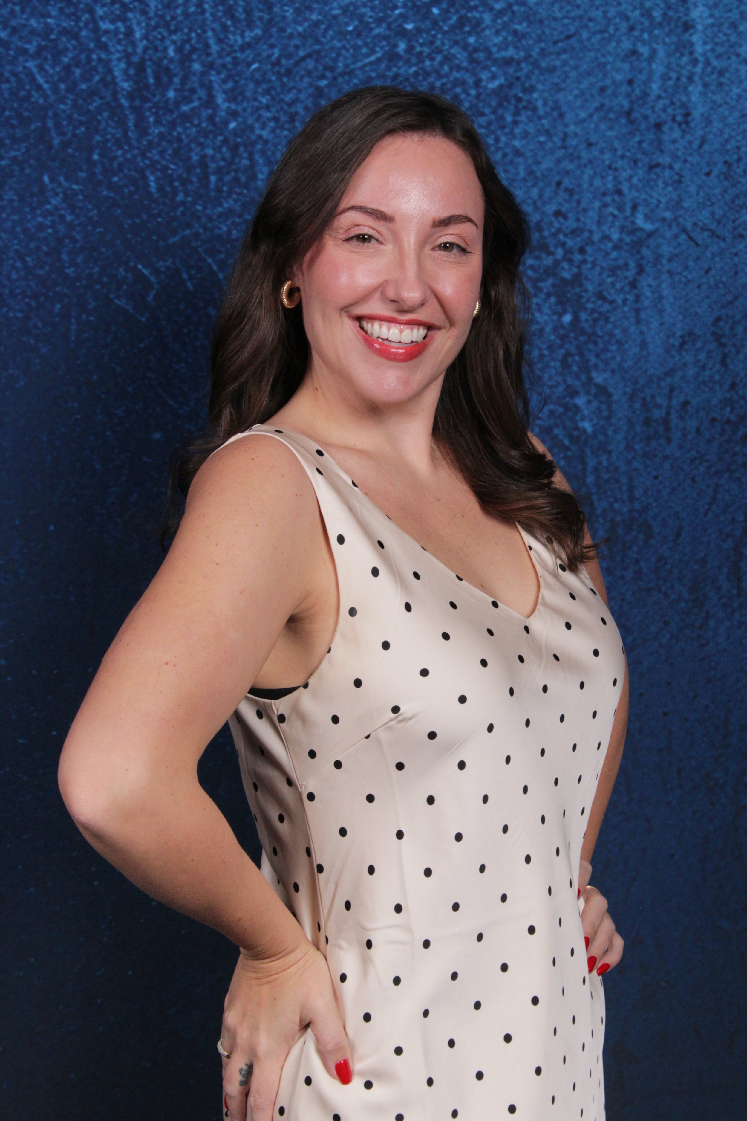 A woman with brown, wavy hair, smiling and wearing a sleeveless white dress with black polka dots, standing against a dark blue textured background.