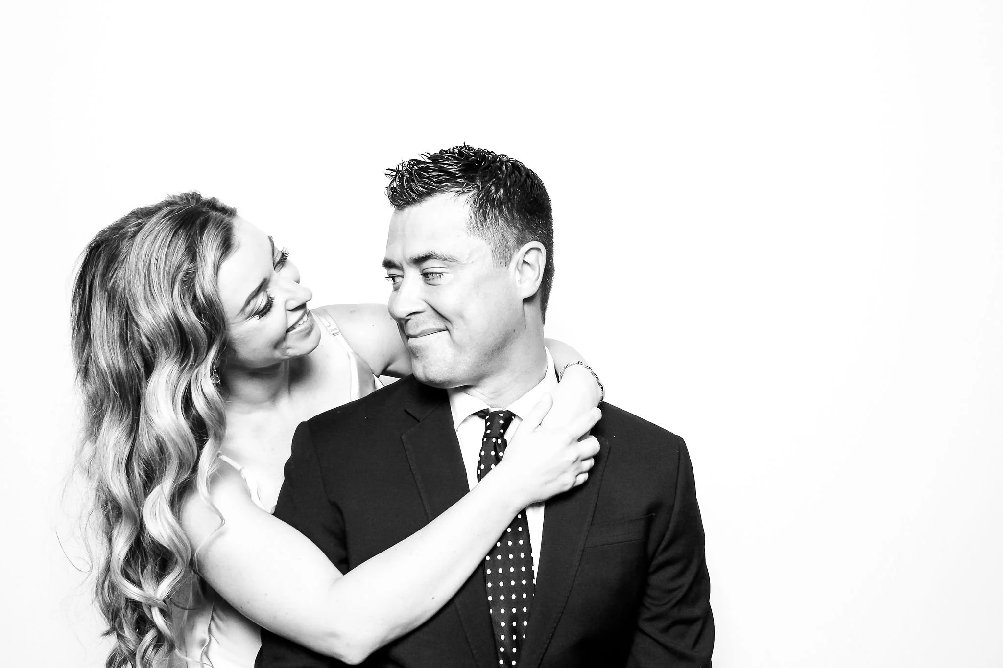A woman with wavy hair smiling and leaning on a man's shoulder, who is in a suit and tie, against a plain white background.