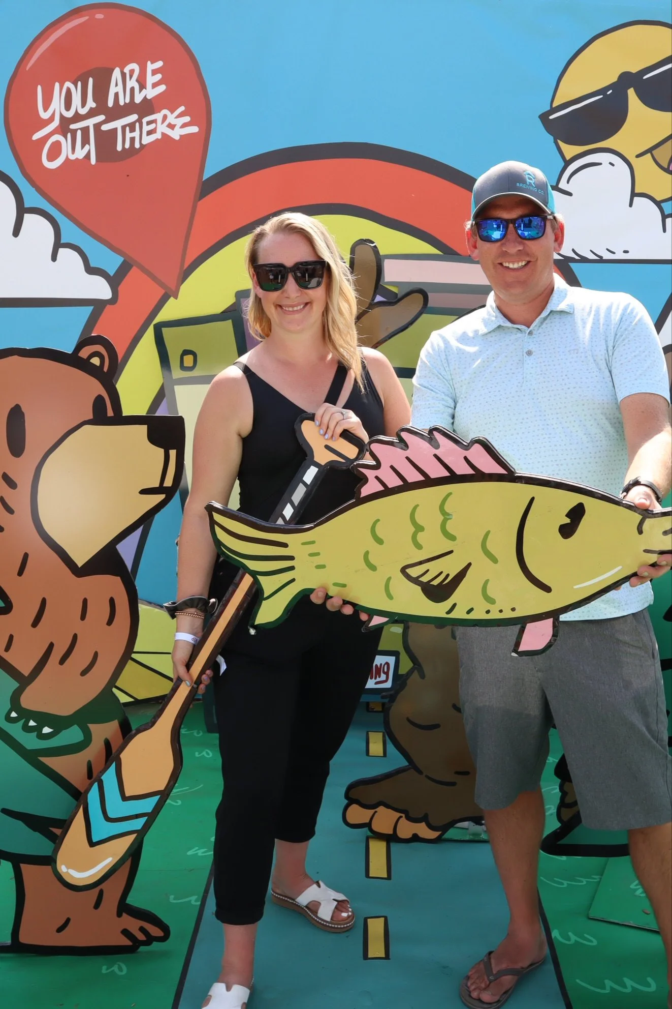 Two people smiling and holding a large cartoon fish cutout, standing in front of a colorful, cartoon-style backdrop with animals and text that reads "You are out there."