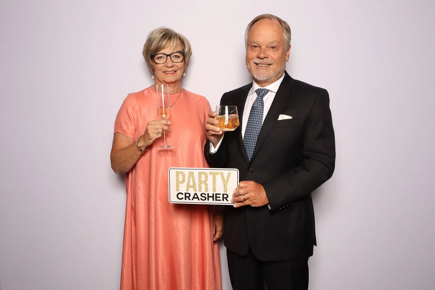 An elderly woman and man in formal attire celebrating, holding drinks and a sign that reads 'Party Crasher', standing against a plain white wall.