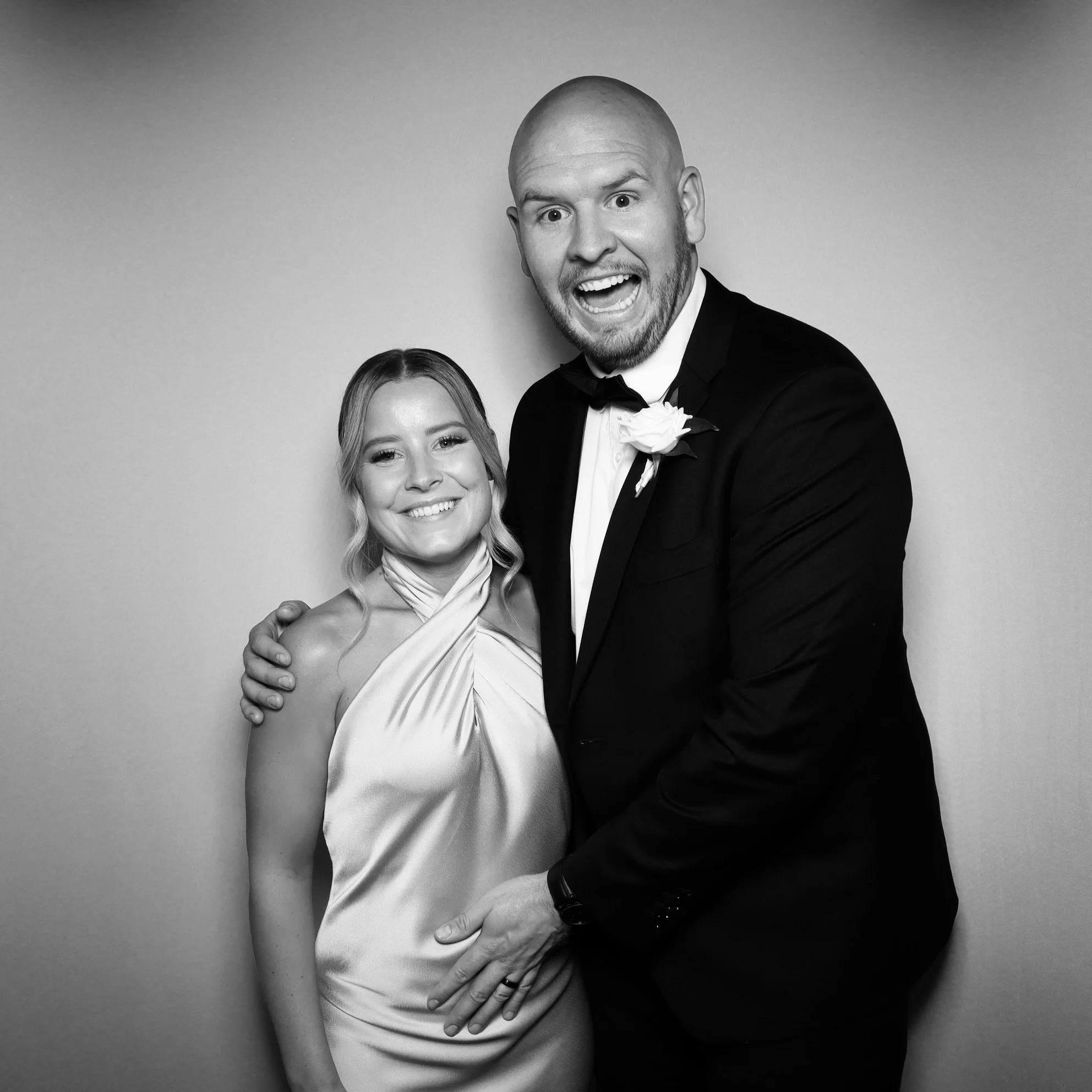 A smiling woman in a satin dress stands next to a man in a tuxedo with a boutonnière, both smiling and posing together against a plain background.