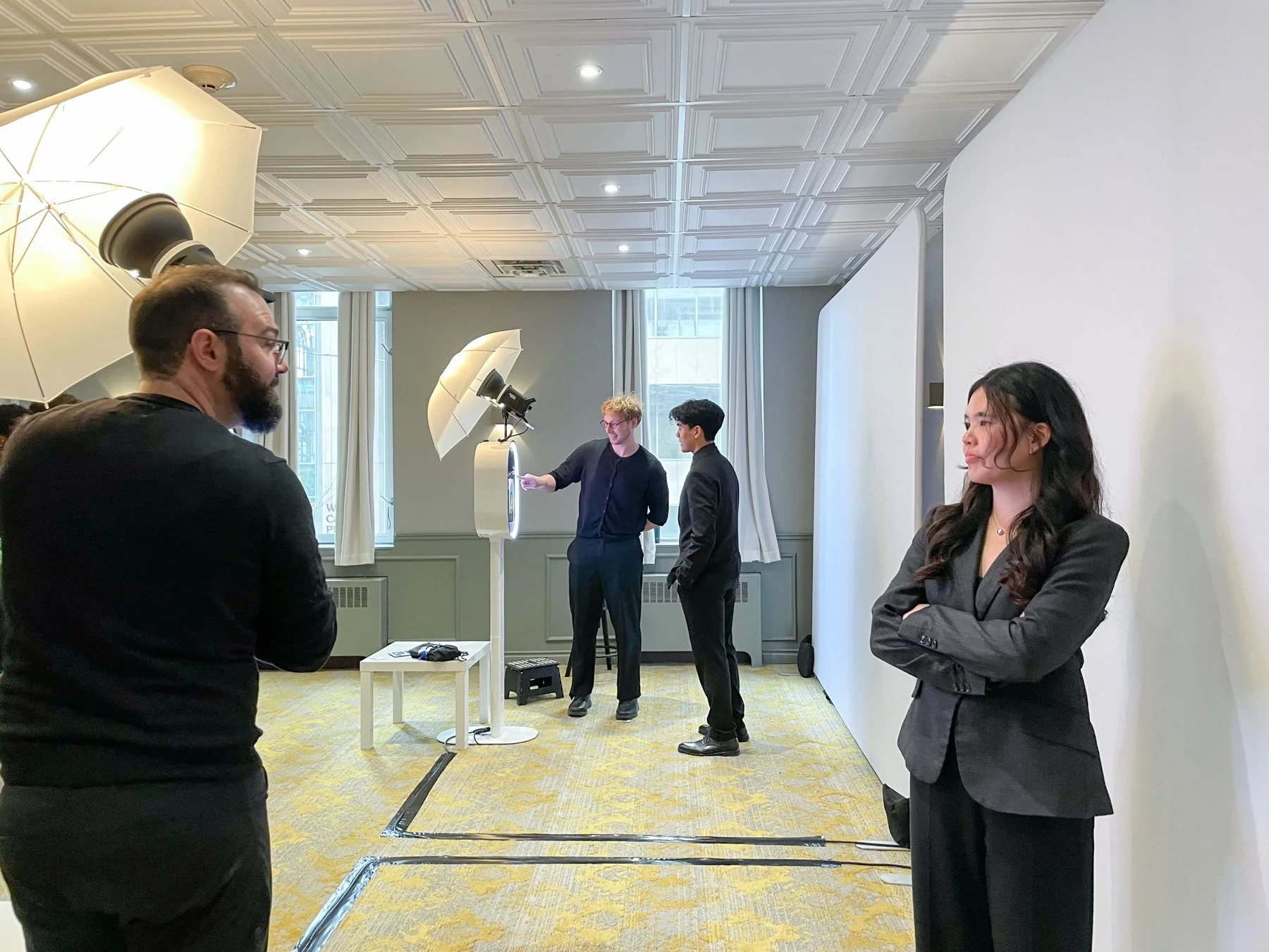 A woman in a black suit standing with arms crossed in front of a white backdrop, with photographers and a man in the background setting up photography equipment in a brightly lit room.