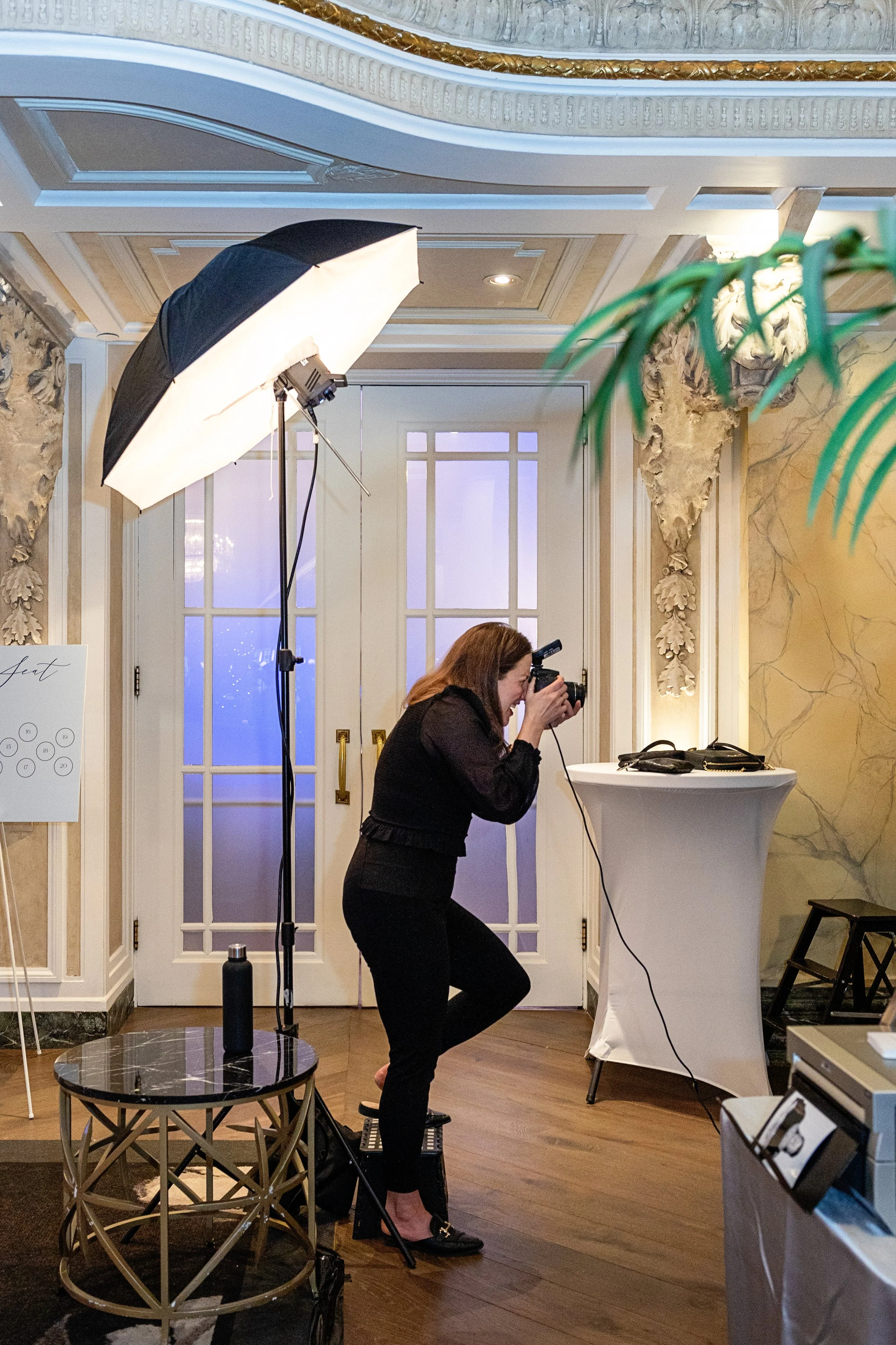 A woman taking a photograph with a camera behind a photography lighting setup in an elegant indoor space.
