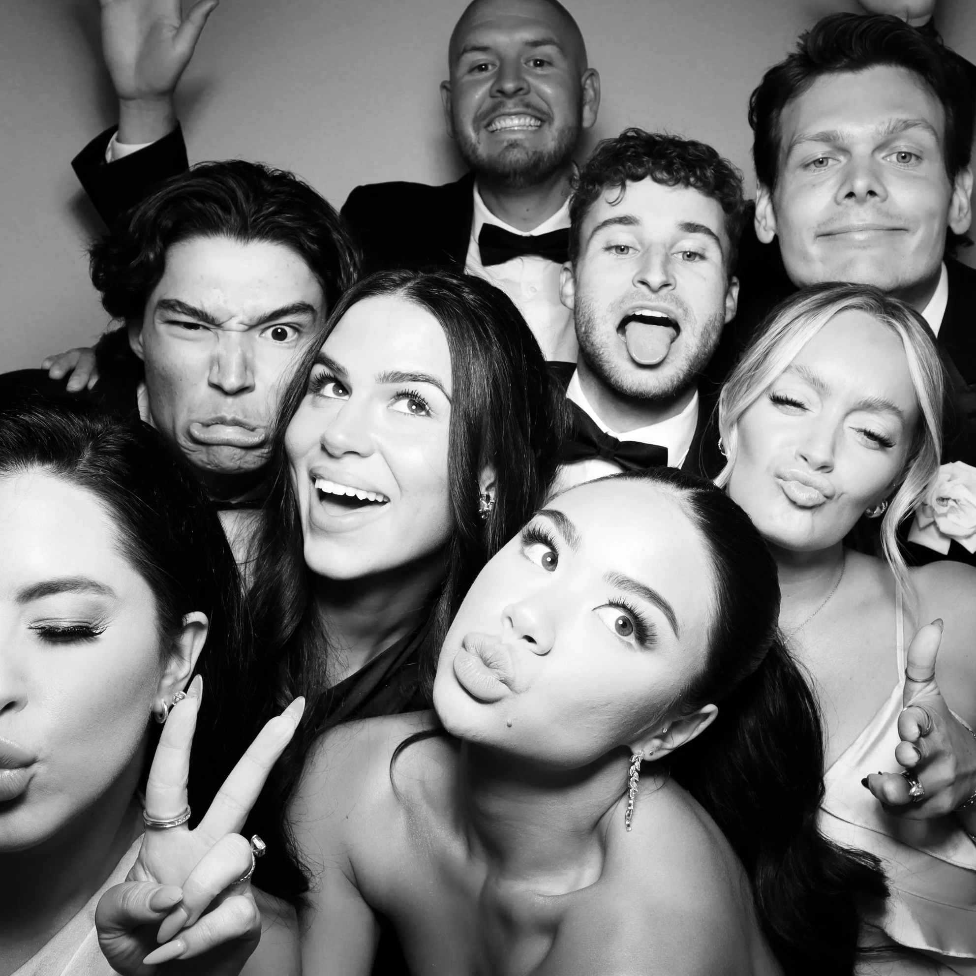 Group of young adults dressed in formal attire taking a playful selfie, making silly faces and posing together.