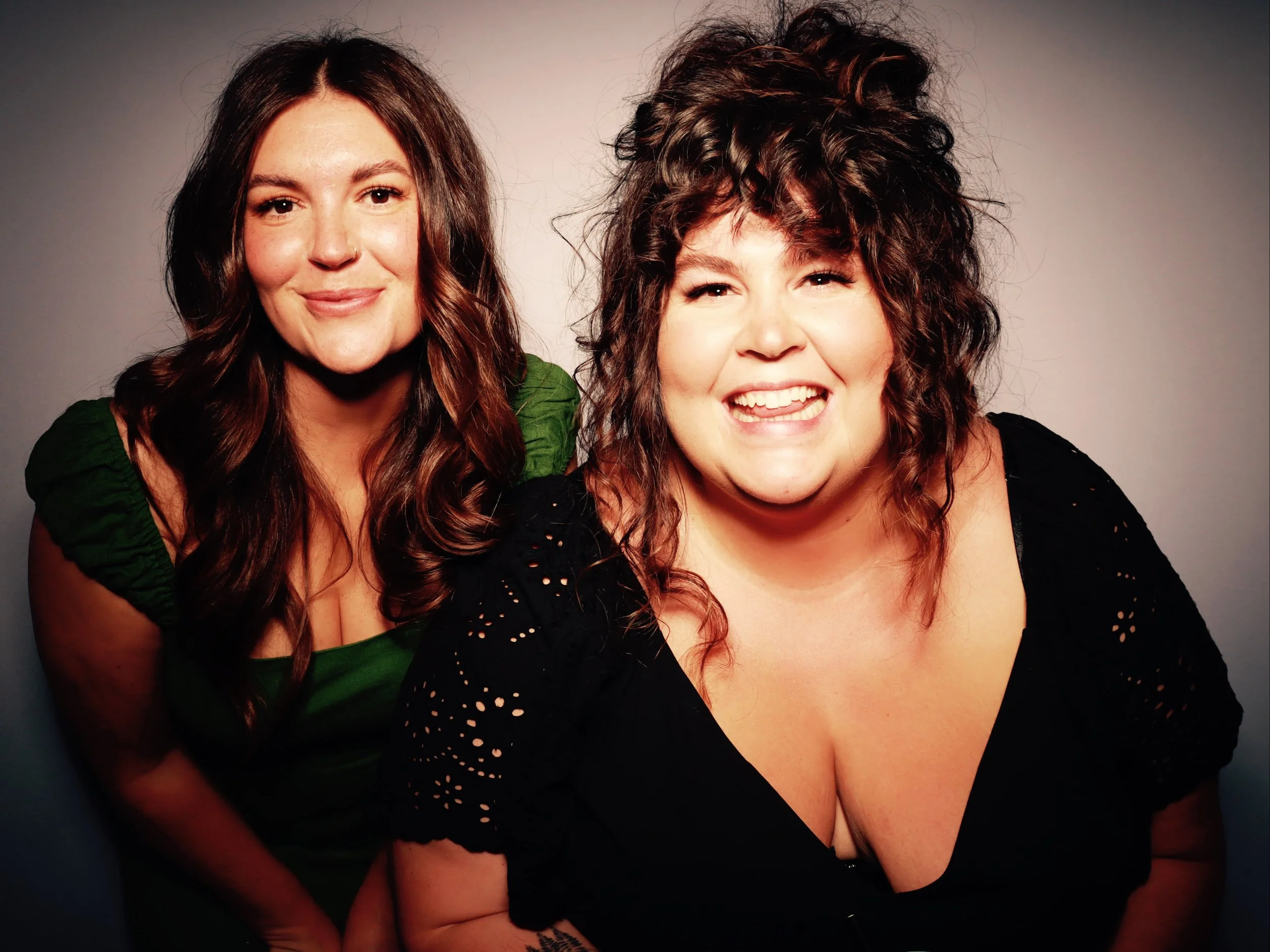 Two women smiling and posing together in a studio with a plain background, one with long brown wavy hair wearing a green top, and the other with curly dark hair wearing a black top with lace details.