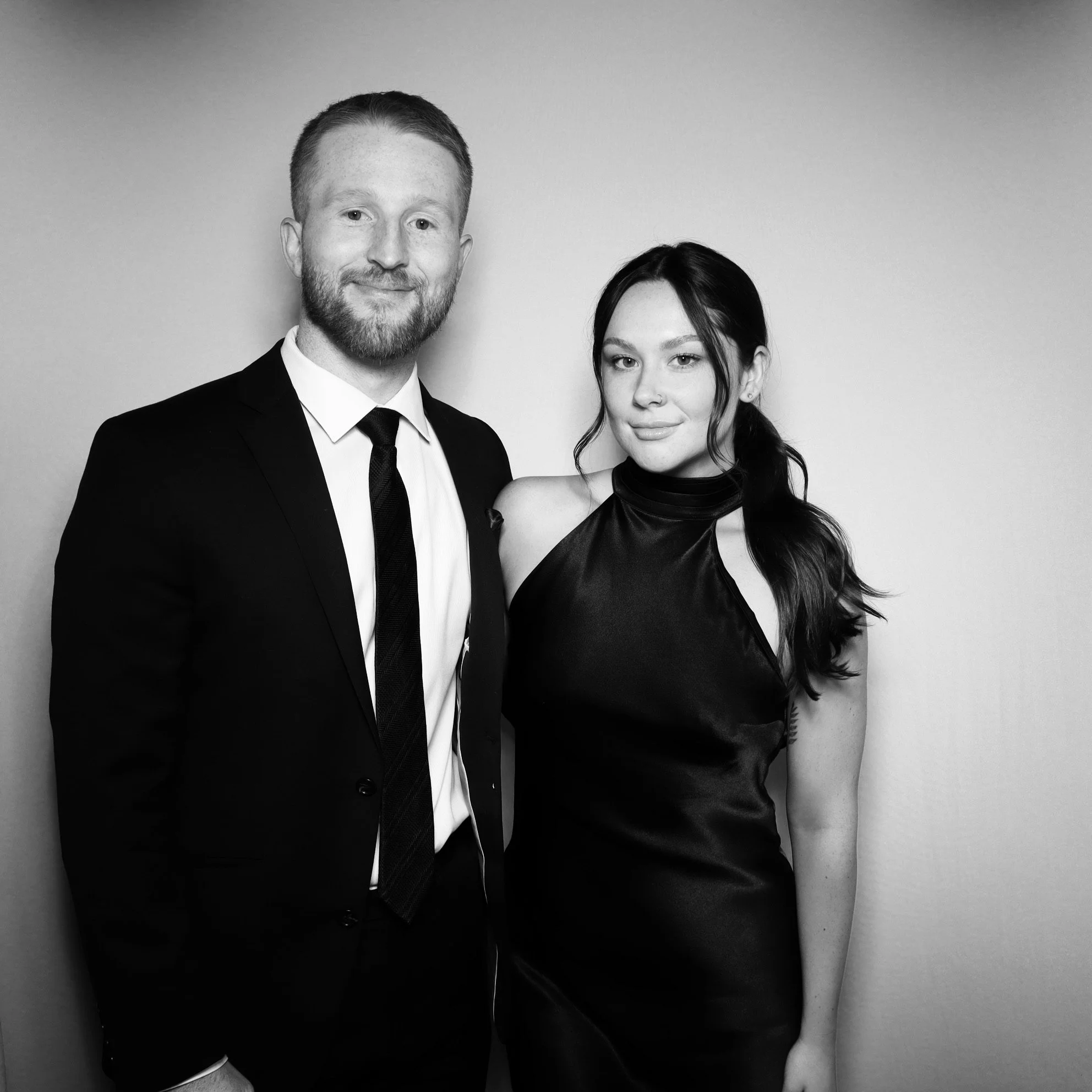 A man in a suit and tie standing next to a woman in a sleeveless silk dress. Both are smiling and looking at the camera, standing against a plain light-colored background.