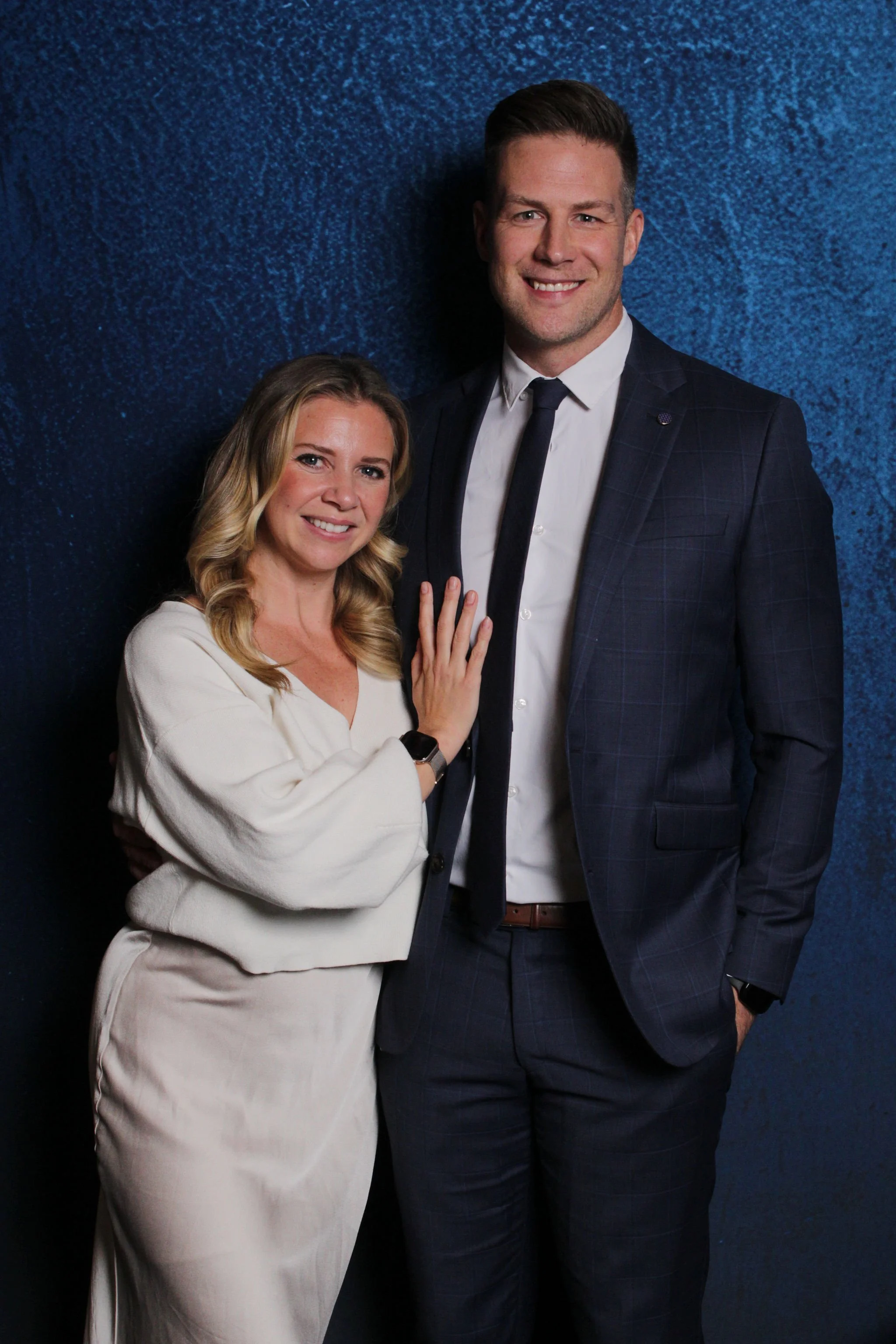 A smiling woman with blonde hair wearing a white dress and a man with short brown hair wearing a dark blue suit, white shirt, and black tie, standing against a textured blue wall.