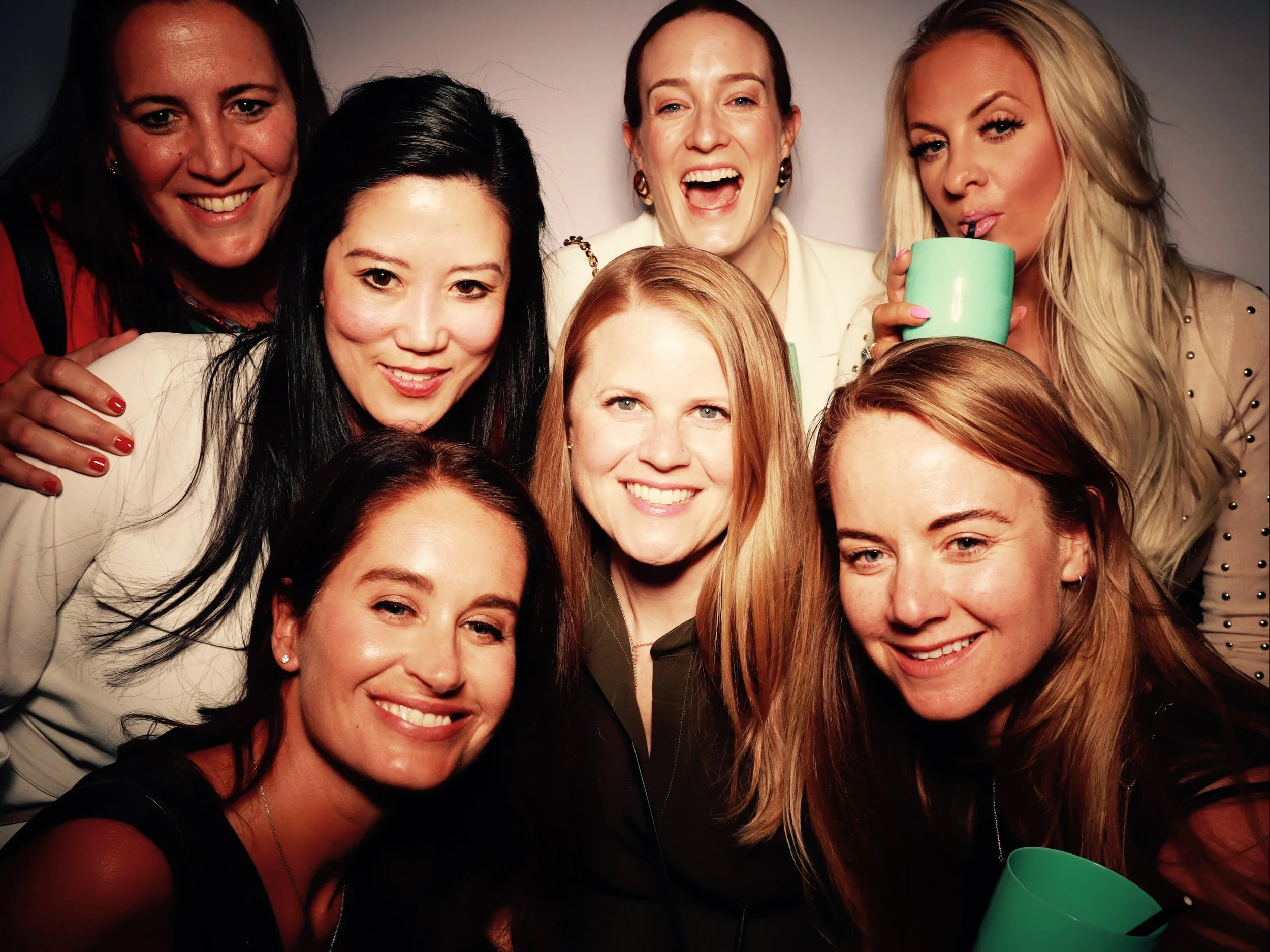Group of nine women smiling and posing for a photo, some holding mugs, in a casual setting.