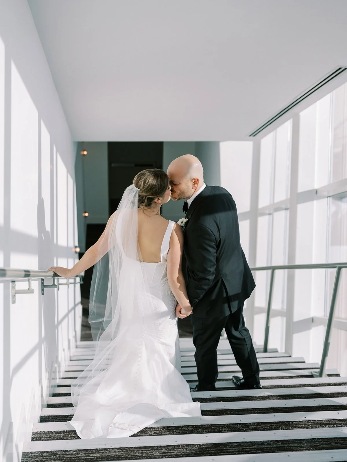 A bride and groom share a kiss on a stairway in a modern, sunlit building.