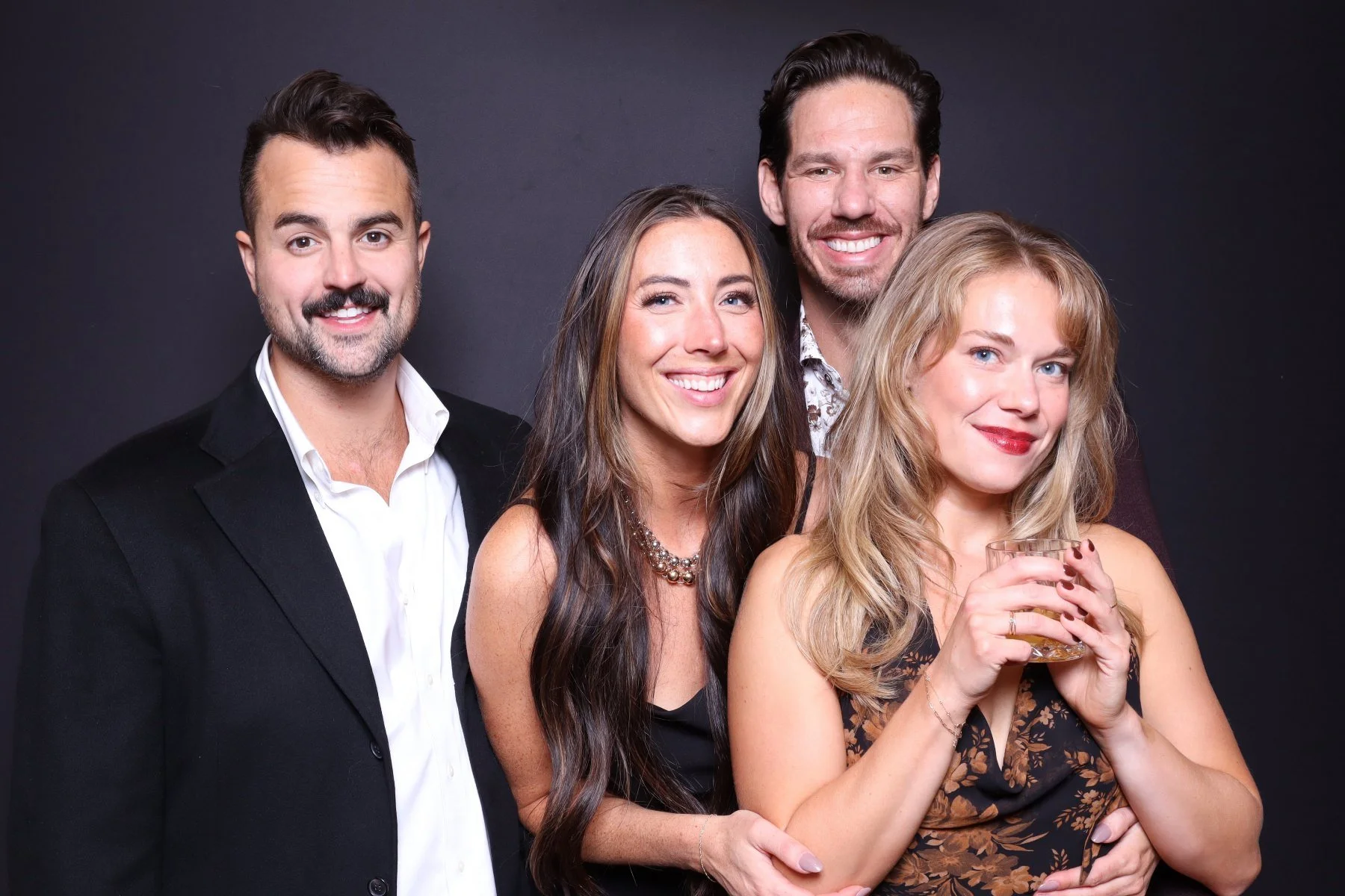 Four adults posing for a photo against a dark background, smiling, with one woman holding a glass of drink.