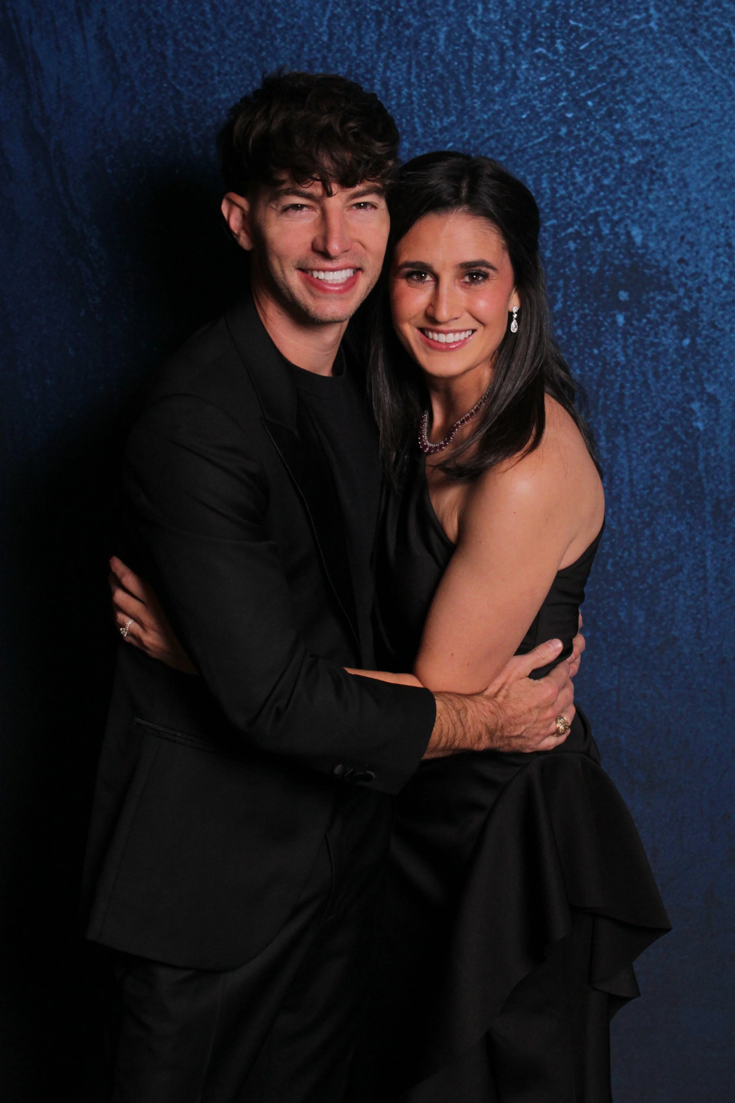 A smiling man and woman in formal attire hugging each other against a blue textured background.