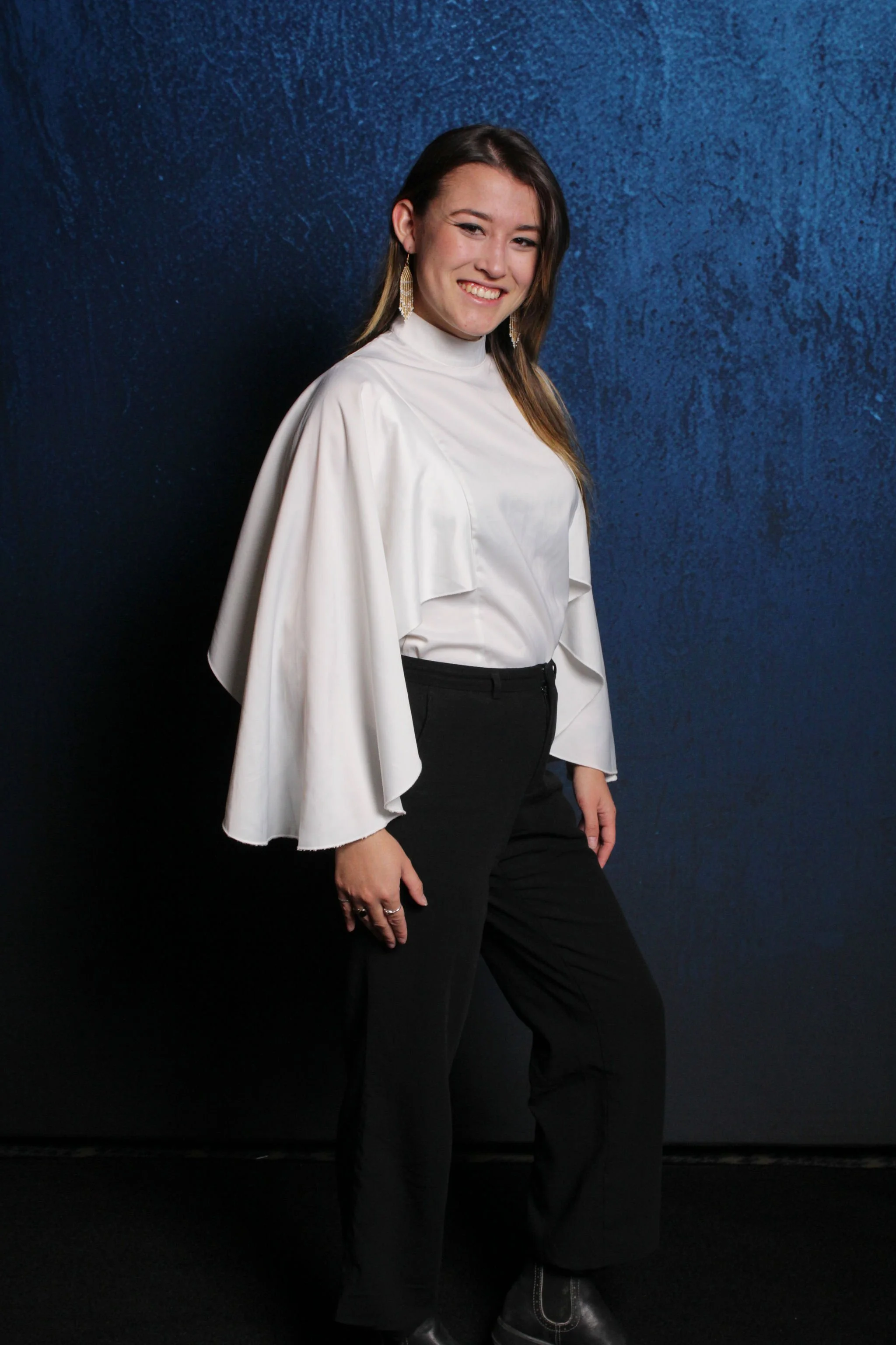 A woman in a white top with cape sleeves and black pants standing against a dark blue textured background, smiling at the camera.
