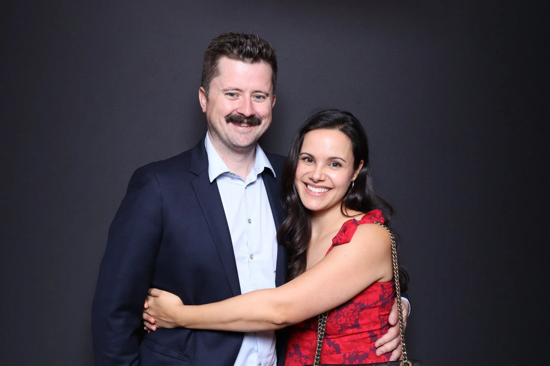 A smiling man in a dark suit and a woman in a red dress with a floral pattern hugging against a dark background.