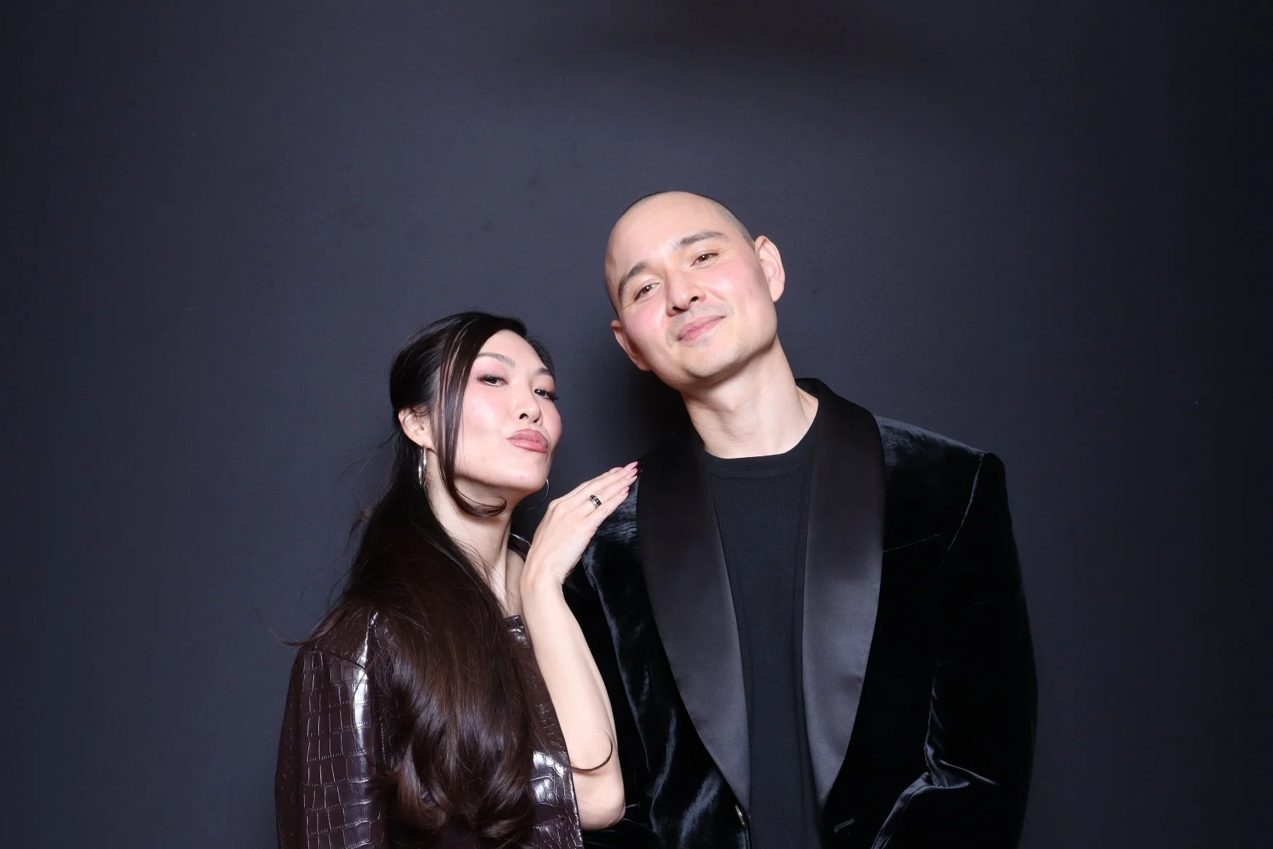 A woman and a man pose together against a black background. The woman has long dark hair and is wearing a dark shiny jacket, while the man is bald and dressed in a black velvet blazer and a black shirt.