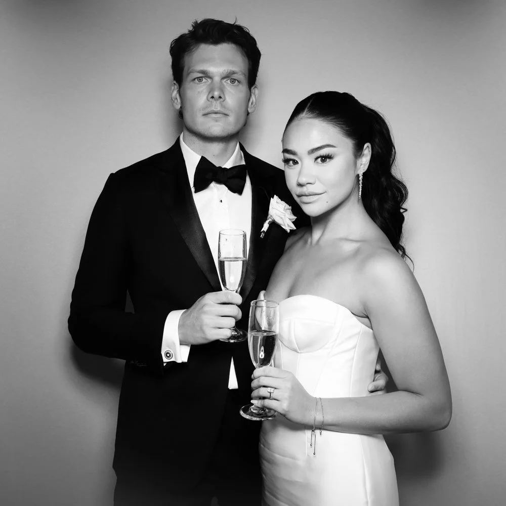 Elegant couple in wedding attire holding glasses of champagne, standing against plain background, monochrome photo.